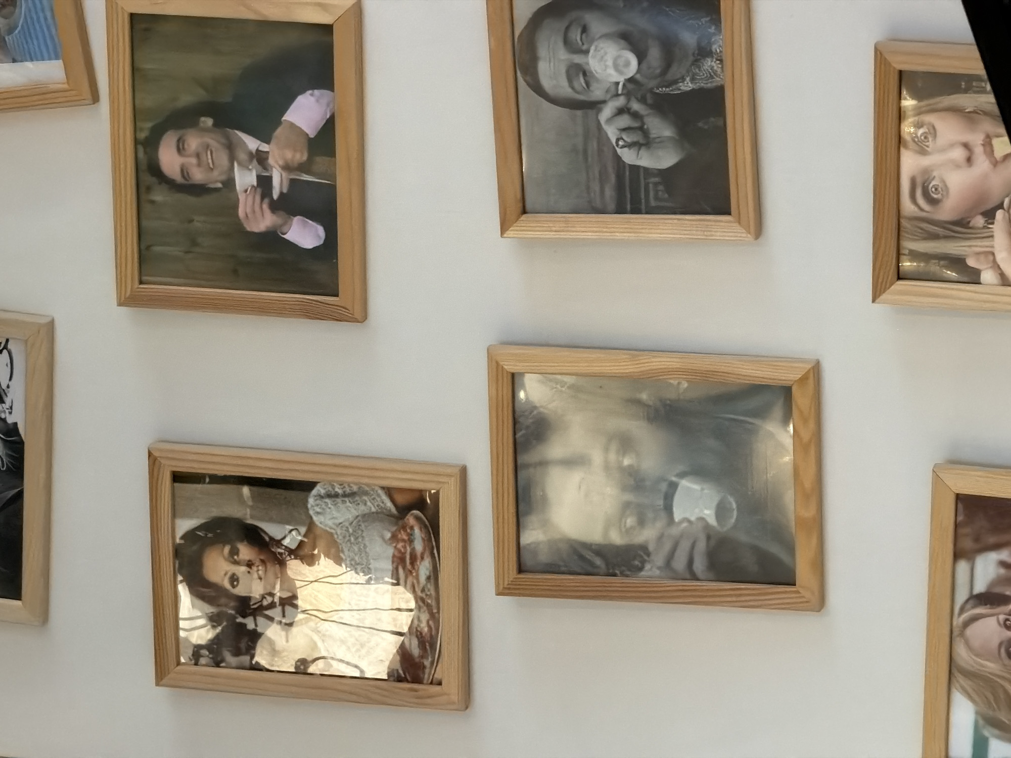 A collection of framed photographs hang on a white wall. The photos, primarily black and white, appear to depict individuals engaged in various activities, such as drinking from a cup or smoking. The frames are light wood. The angle and composition of the photo suggest an interior setting. The location of the image is unknown, and there is no information to indicate it is in Izhevsk, Russia.