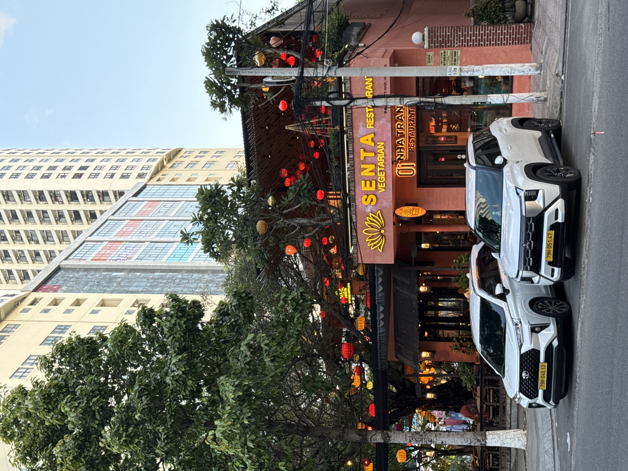 The image shows the exterior of the "Senta Vegetarian Restaurant" in Nha Trang, Vietnam, during the daytime. The restaurant has a vibrant pink facade adorned with neon signs and hanging red lanterns. Two white SUVs are parked on the street in front of the establishment. The background features a tall, modern building with a light-colored exterior and multiple windows, characteristic of urban architecture in Nha Trang. Trees with lush green foliage frame the scene, and the sky is a clear, light blue. The street appears to be a typical urban thoroughfare, with a sidewalk and utility poles visible. The overall atmosphere is that of a bustling city setting with a focus on the restaurant and its surroundings.