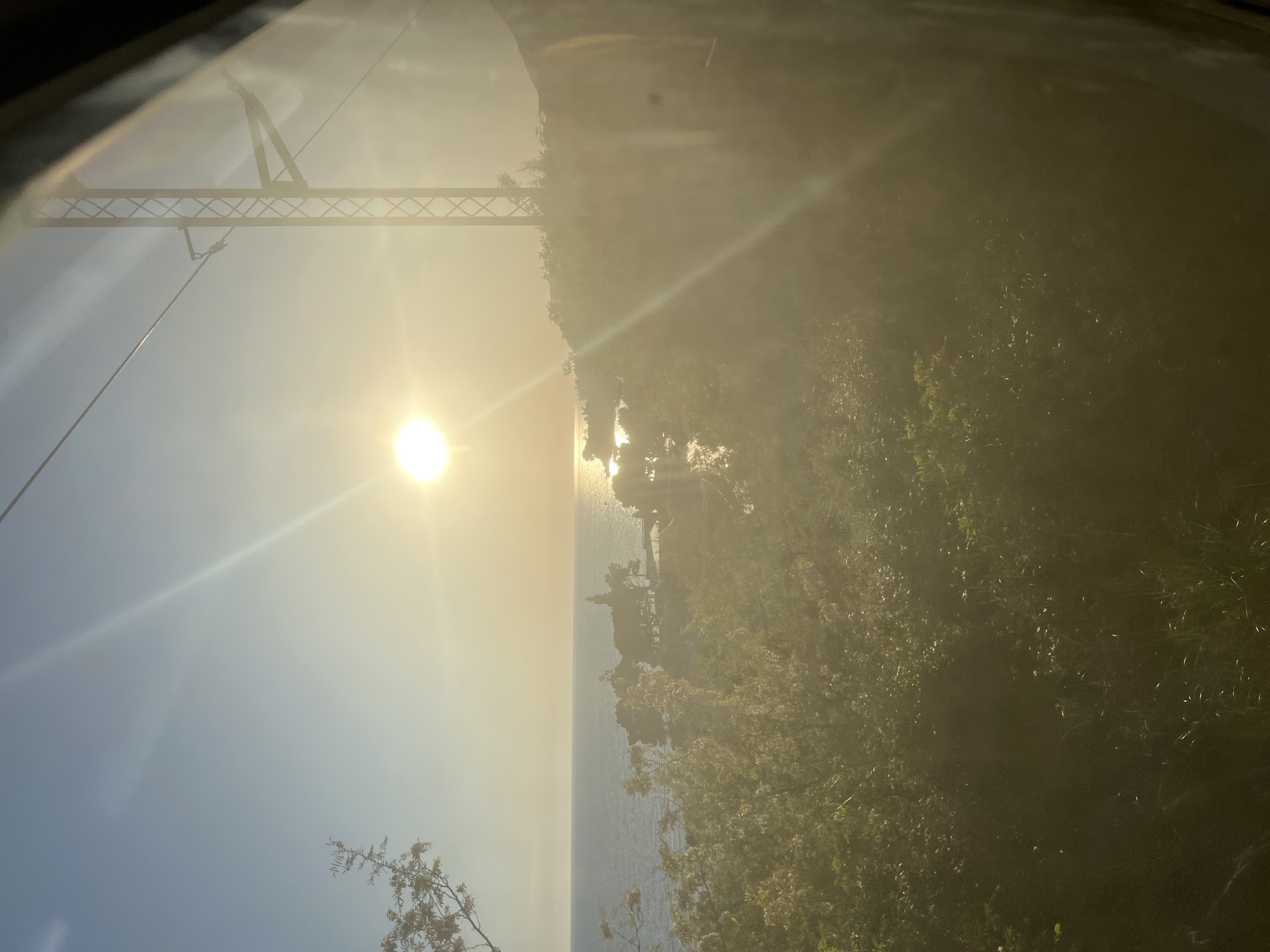 The image shows a landscape view with a bright sun obscuring many details. To the left of the image is a bridge structure, seen from below. Adjacent to the bridge are water and a coastline with buildings. The right side of the frame is dense vegetation. The location is likely near Bar, Montenegro, given the geographical features. No people are visible.