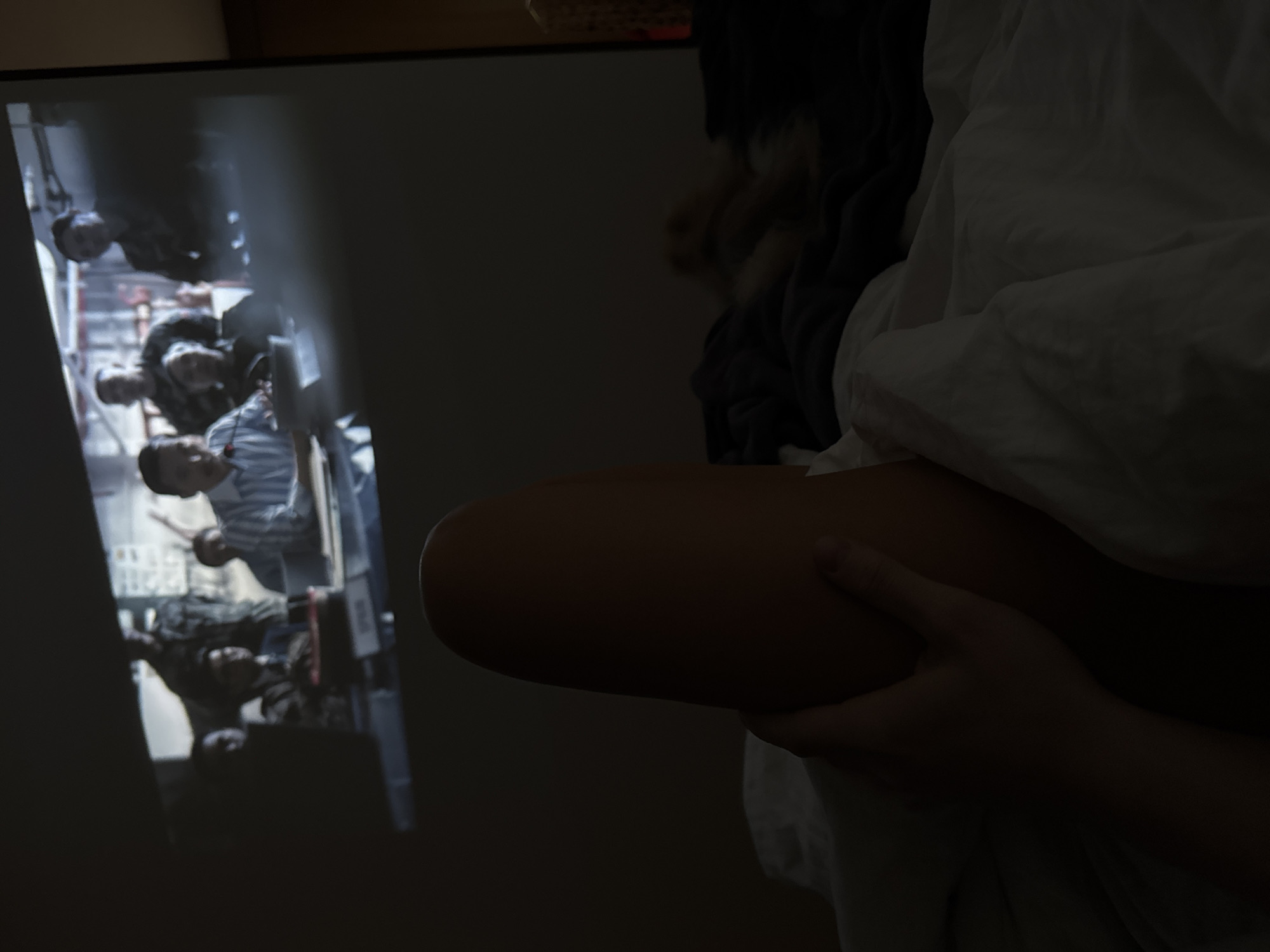 A person sits in bed with their leg propped up, clutching it with their hand. To their left, a projected image shows a group of people standing in what appears to be an elevator. The location is indeterminate.
