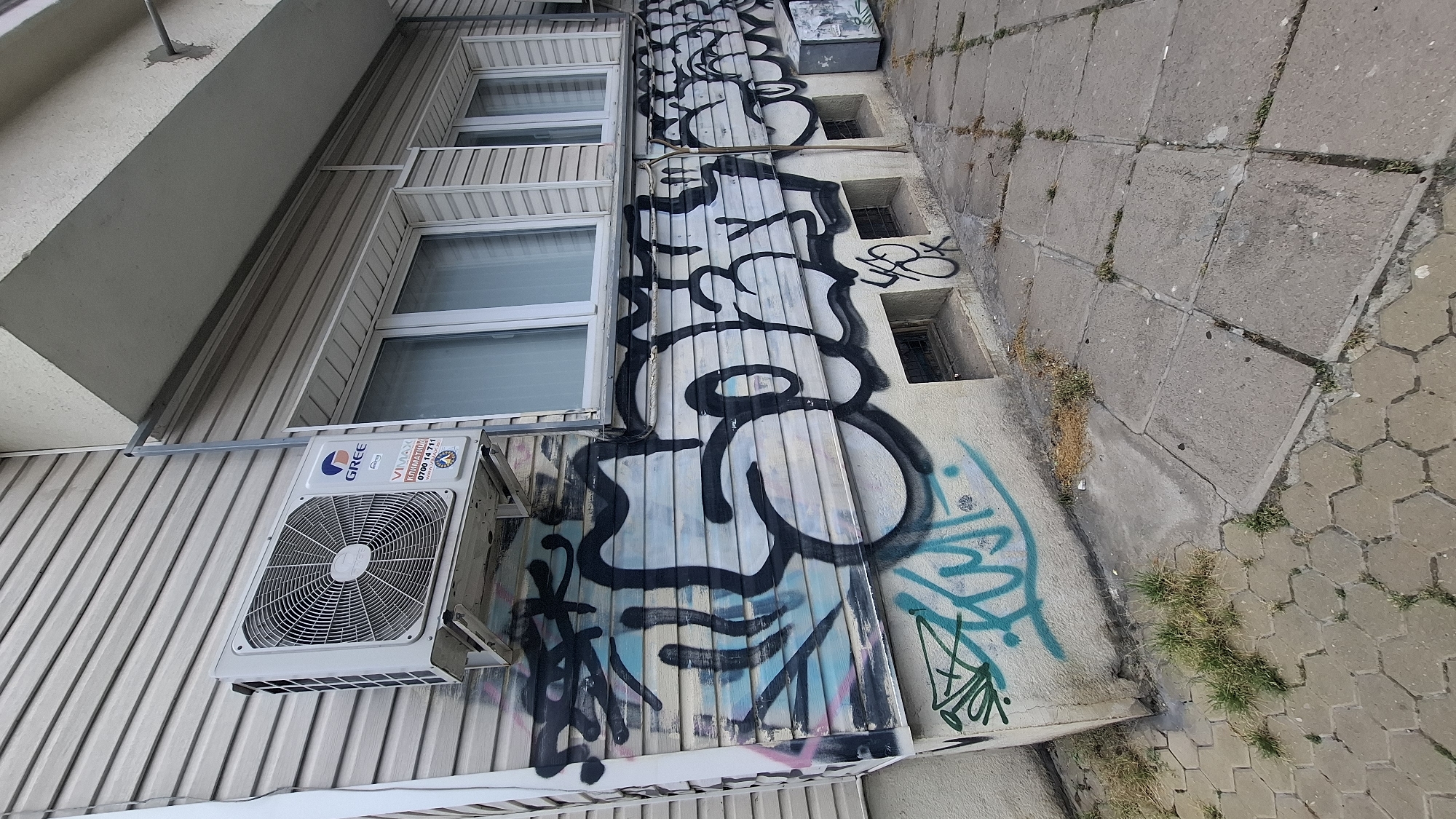 An outdoor shot of a building's exterior in Sofia, Bulgaria, features an air conditioning unit mounted on a light-colored, horizontal-paneled wall. Below the unit, the wall is heavily graffitied with large, black, stylized lettering, some outlined in pink and light blue, and a smaller teal tag with a starburst shape. The building has several large windows with light-colored blinds. A concrete sidewalk with hexagonal and square paving stones runs along the base of the wall, with patches of grass and dry vegetation growing in cracks and crevices. Small, recessed window wells with metal grates are visible at the bottom of the building. The overall scene depicts an urban environment with signs of wear and artistic expression.