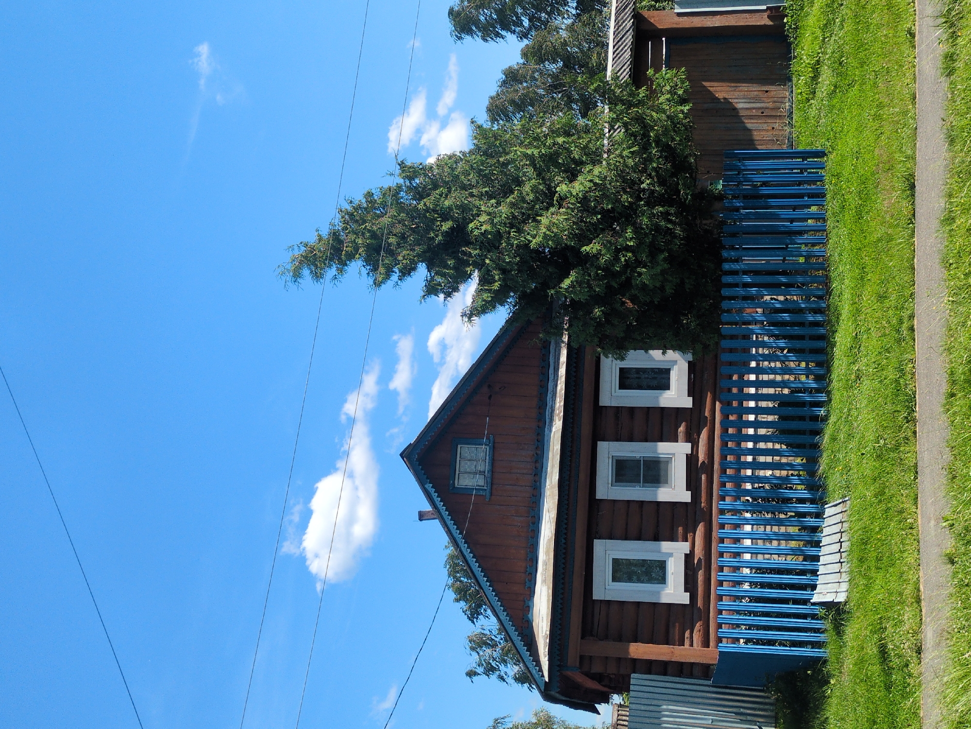 This image shows a rustic wooden house in a village setting. The house has a steeply pitched roof with decorative trim and multiple windows. A bright blue fence runs along the side of the house, bordering a lush green lawn and a paved path. A tall evergreen tree is situated next to the house, its branches reaching towards the bright blue sky scattered with fluffy white clouds. Power lines are visible against the sky, suggesting the presence of modern infrastructure in this otherwise traditional environment. The sun appears to be shining brightly, indicating a clear, warm day. The overall impression is one of peaceful rural life in Velet'ma, Russia, with a blend of old-fashioned charm and everyday practicality. There is no visible text in the image.