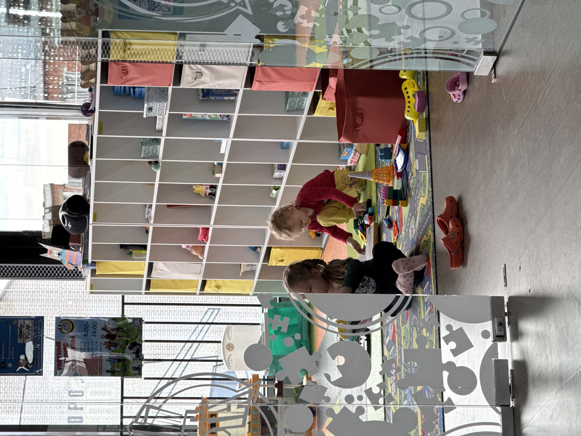 Here is a detailed description of the image:
**Setting:**
The image appears to be taken inside a children's playroom, possibly in a shopping center or public space in Votkinsk, Russia. This is inferred from the Russian text on the promotional posters visible through the glass partition.
**People:**
There are two children in the scene, likely girls. One is wearing a red shirt and yellow pants and is interacting with toys on a colorful rug. The other is wearing a dark shirt and is also playing with the toys.
**Objects:**
* **Shelves:** A large white shelving unit is filled with various toys and containers.
* **Toys:** A colorful rug is covered with various toys.
* **Clothing:** Clothes and shoes are placed near the rug.
* **Furniture**: Tables and chairs appear to be set up.
* **Promotional posters:** Posters are visible, advertising children's activities such as a disco with cotton candy, alongside contact information including a phone number and business hours.
* **Glass partition:** A glass partition with playful designs featuring puzzle pieces and circles separates the play area from the other side.
**Actions/Interactions:**
The children are actively engaged in playing with the toys on the rug.
**Environment:**
The room seems to be a dedicated play area with ample toys and shelves for storage. The environment appears child-friendly, aiming to be a playful and stimulating space. The presence of the glass partition suggests a level of supervision or separation from other parts of the building. The presence of the posters point to it being a public area and recreational place.