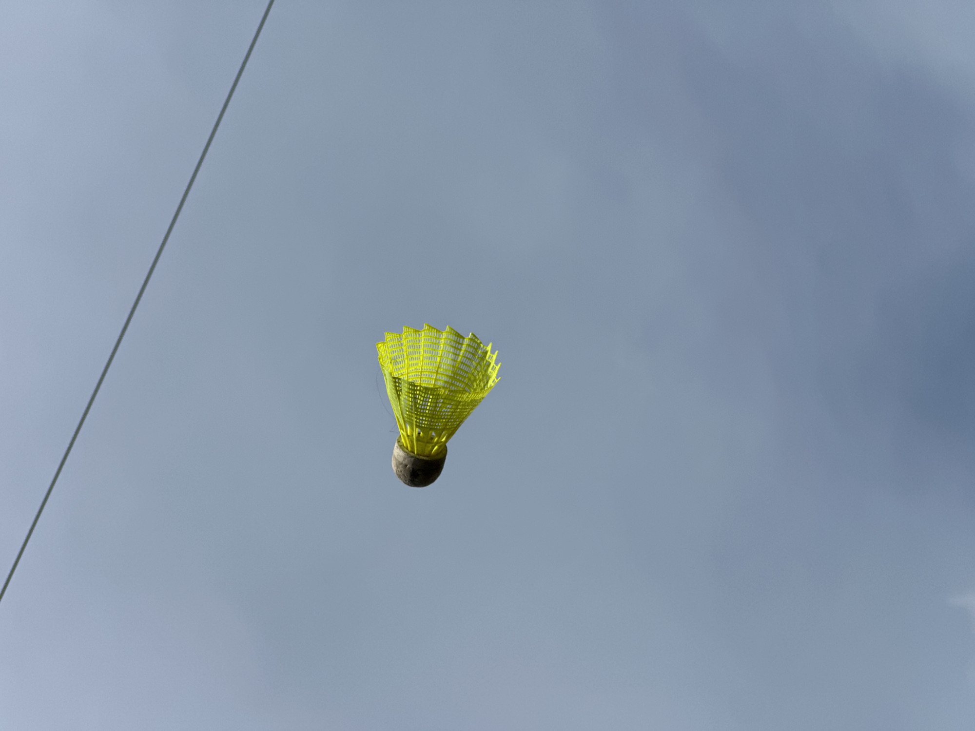 A yellow badminton shuttlecock is suspended in mid-air against a backdrop of a cloudy, light blue sky. A thin wire diagonally crosses the upper left portion of the frame. There are no discernible buildings or other identifying features visible that would indicate the location is Nizhny Novgorod, Russia. There are no people present in the image.