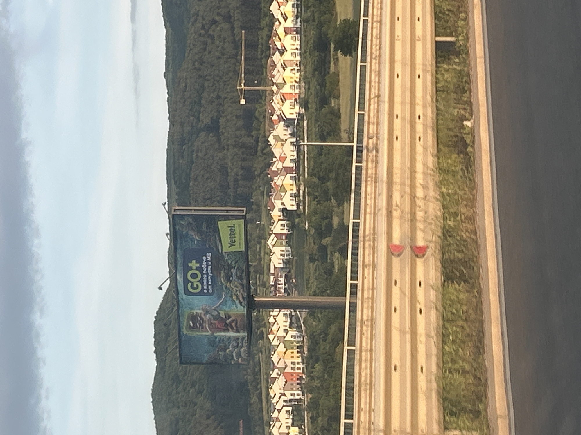 The image depicts a highway with a billboard, houses, and a forest backdrop. The billboard promotes "GO+" and "Yettel," suggesting a telecommunications advertisement. The houses appear to be closely packed together, possibly a residential development. A construction crane is visible among the houses. The forest creates a natural barrier behind the houses. The highway runs along the right side of the image. The sky is overcast. The location appears to be the city of Unknown, Unknown.