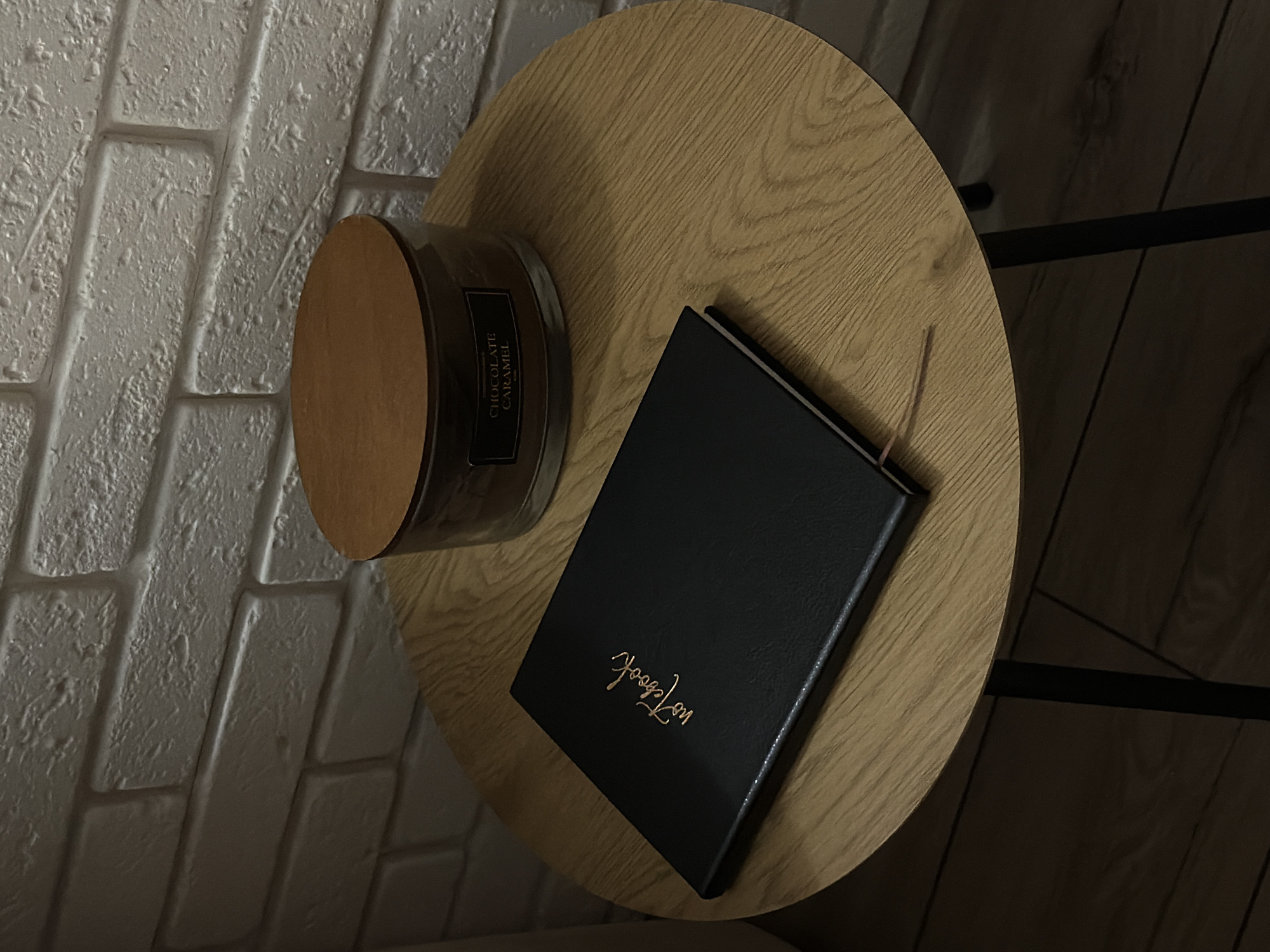 A high-angle shot reveals a small, round wooden side table placed against a white brick wall. On the table, there is a candle labeled "CHOCOLATE CARAMEL" with a wooden lid and a black notebook with the word "Notebook" inscribed in gold lettering. The table has a black metal leg that is partially visible. The environment appears to be indoors, potentially in a residential setting.