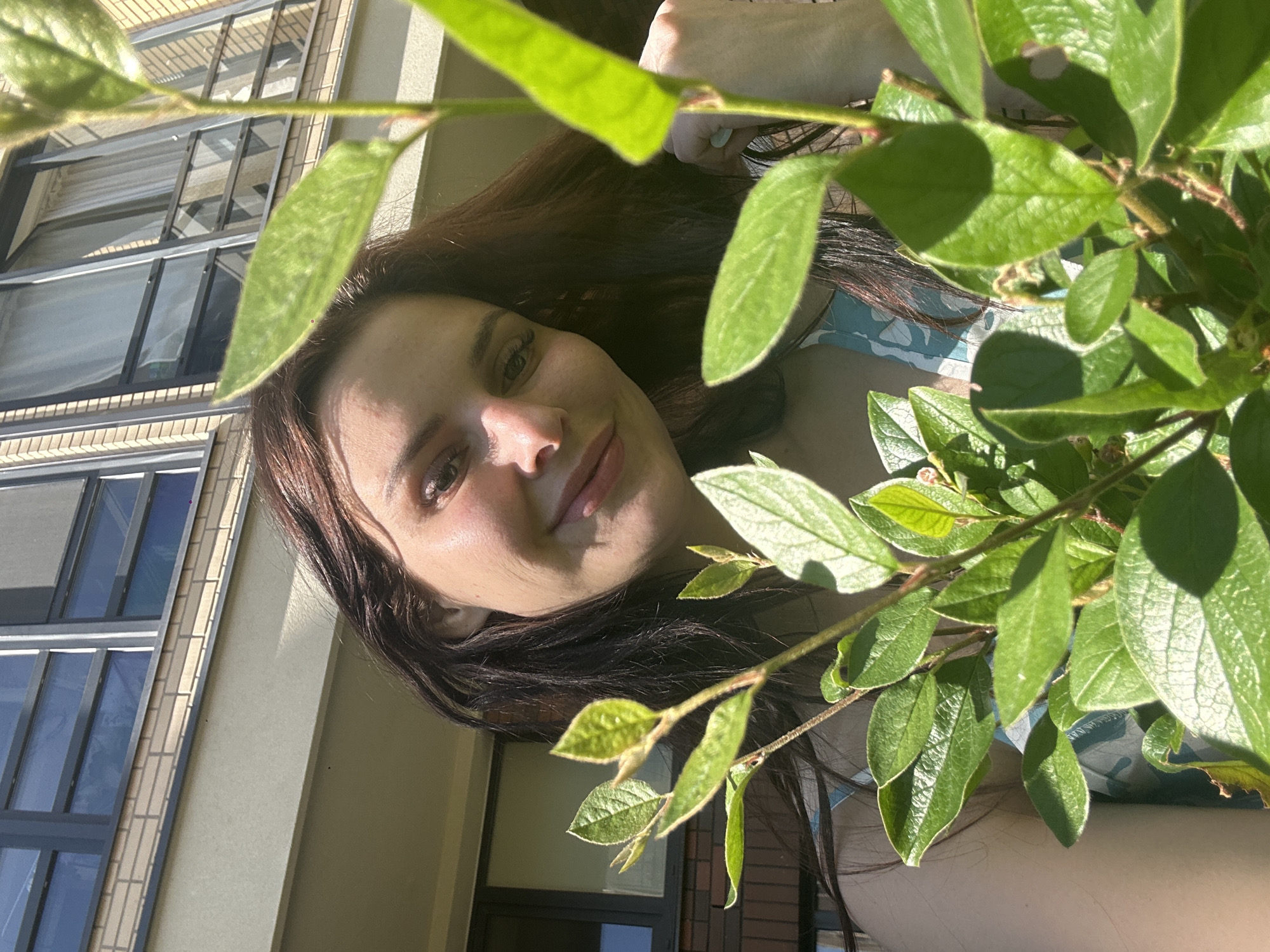 In an outdoor urban setting, a woman with dark hair and fair skin is captured in a low-angle shot. She is looking towards the camera with a gentle smile, her face illuminated by sunlight. She appears to be wearing a light-colored top with a blue and white pattern. In the foreground, lush green foliage from a plant partially obscures the woman and the background. Behind her, a building with multiple windows and beige brickwork is visible, suggesting a city environment. The lighting indicates it is daytime.