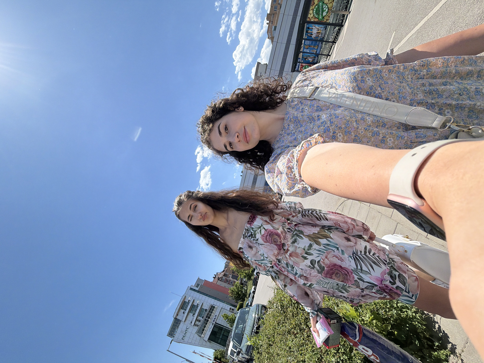 Two young women are taking a selfie outdoors in Sofia, Bulgaria. The woman on the left is wearing a floral off-the-shoulder dress and has her hair down. She is holding a package in her left hand and is making a face at the camera. The woman on the right is wearing a floral dress and has curly hair. She is looking at the camera with a slight smile. They are standing on a sidewalk next to a row of bushes. In the background, there is a modern building with the name "Pamyc" on it, a street with cars, and what appears to be a shopping center with large posters displayed. The sky is blue with some clouds and the sun is shining brightly, casting a lens flare.
