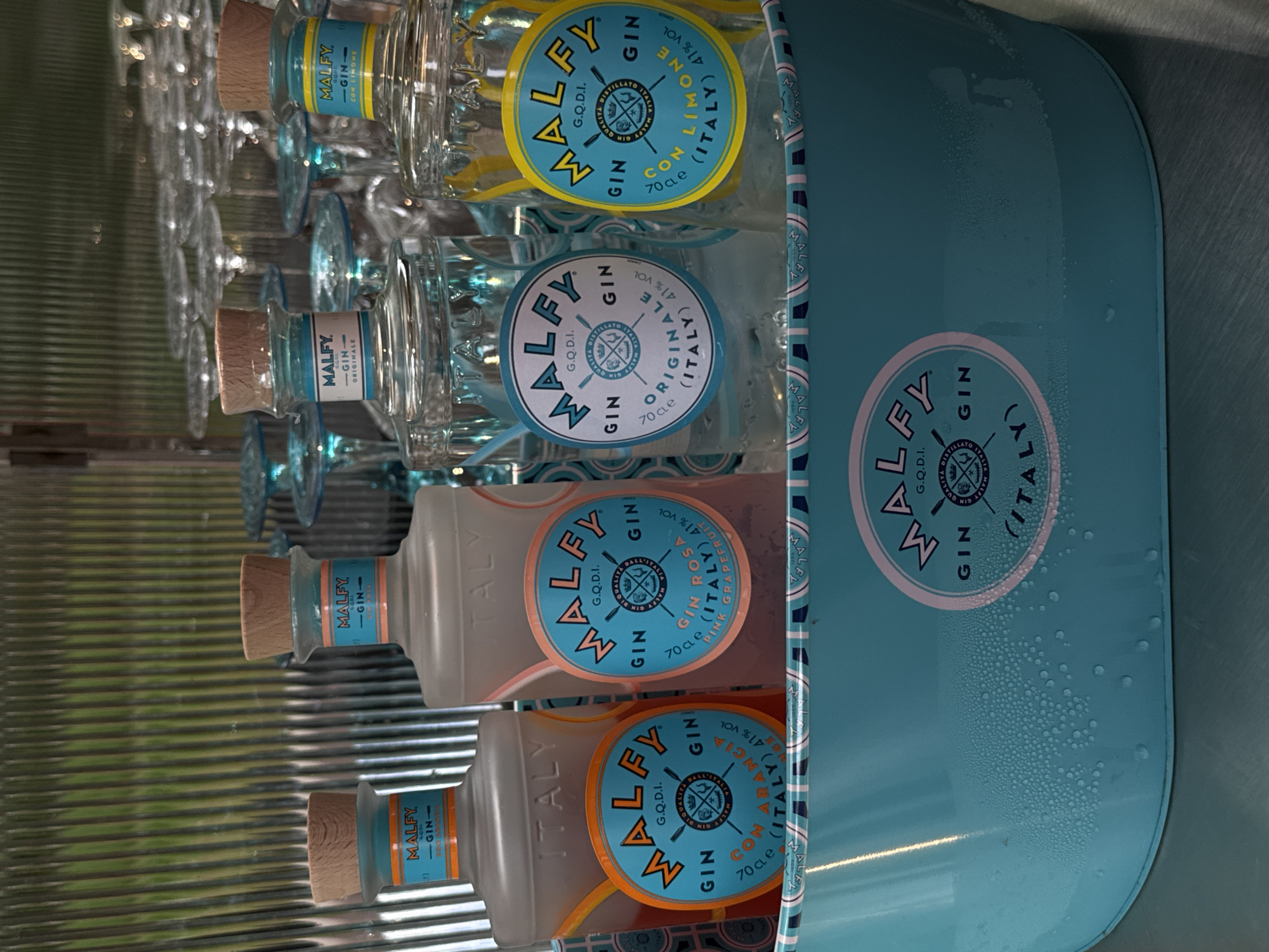 The image shows a collection of Malfy Gin bottles stored in a light blue cooler. The bottles are of different varieties, indicated by the colored labels: lemon, orange, grapefruit, and original. Each bottle has a wooden cap and a light blue label with the words "Malfy Gin." The cooler also has a Malfy Gin label printed on its side. The background is out of focus, but appears to be a metallic surface with horizontal striations. There is no information about Yerevan, Armenia or people in the image.