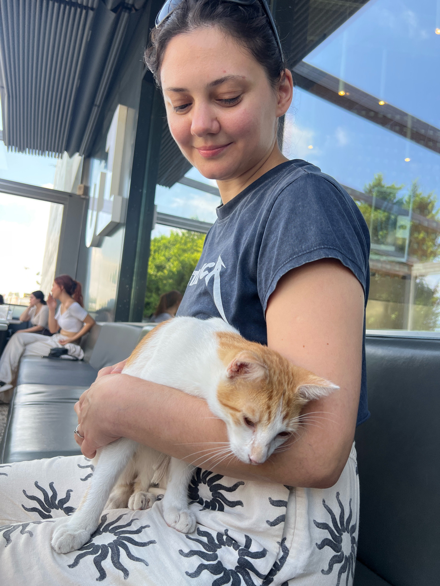 A woman is holding a ginger and white cat in her arms. She is wearing a dark t-shirt and patterned pants. The cat is looking down and appears to be relaxed. In the background, there are other people sitting on couches, a large window with a view of trees and sky, and a modern ceiling structure. The image appears to have been taken indoors at a cafe or similar establishment. The overall lighting suggests it is daytime.