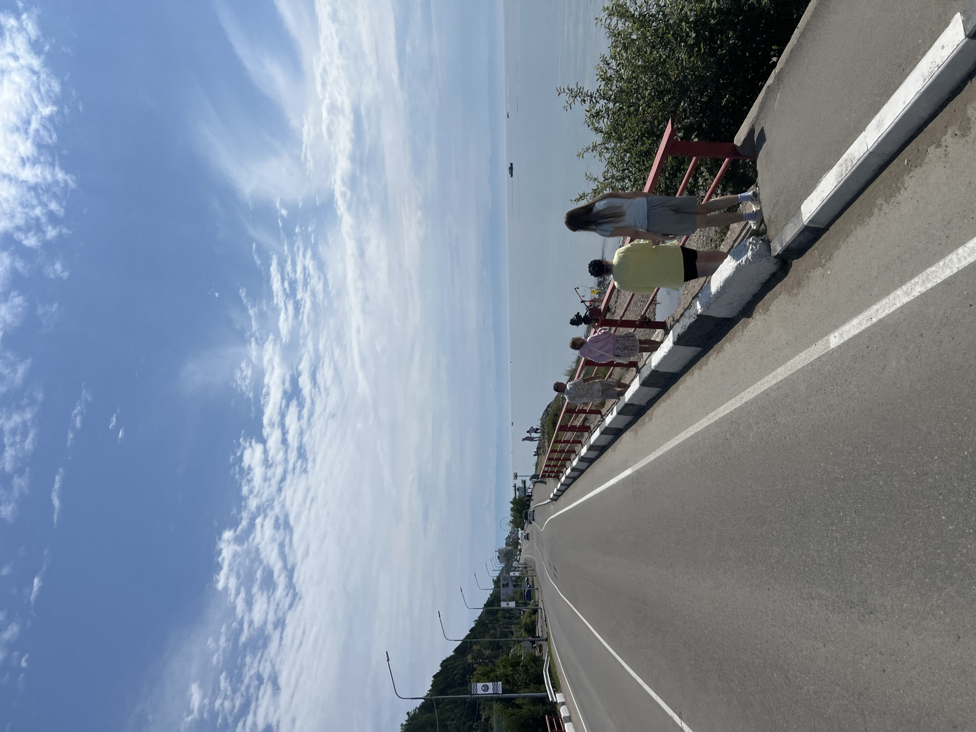 This is a photograph taken from a road that runs alongside a large body of water, likely Lake Baikal, in Listvyanka, Russia. In the foreground, a paved road with white lane markings stretches into the distance. To the right of the road, a sidewalk or pedestrian walkway is bordered by a red railing. Several people are walking or standing on this walkway, facing the water. A few of these individuals are visible in detail, including a woman in a yellow shirt and black shorts, and another woman in a light blue t-shirt and grey shorts. In the background, the road curves, with streetlights lining its side. Further in the distance, a Ferris wheel is visible, suggesting a recreational area. The water is calm and extends to the horizon, where a hazy shoreline is barely discernible. The sky is a clear blue, with scattered white clouds. Small boats are present on the water in the distance. Lush green trees and vegetation can be seen on the hillside to the left of the road. The overall scene depicts a scenic road and promenade area in a coastal town.