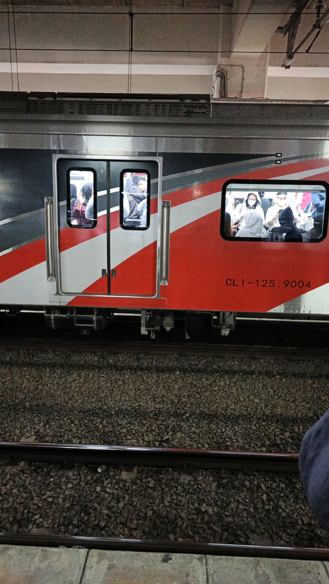 The image shows a side view of a train carriage at a station in South Jakarta, Indonesia. The train has a red and silver livery with a diagonal red stripe. There are windows and doors on the carriage. Through the windows, several passengers are visible, some wearing face masks. One window shows a person in a hijab and another shows a person with a head covering. The train is on tracks, with gravel and concrete visible on the ground. The lighting suggests it is either early morning, late evening, or indoors. The text "CLI-125.9004" is visible on the side of the train.