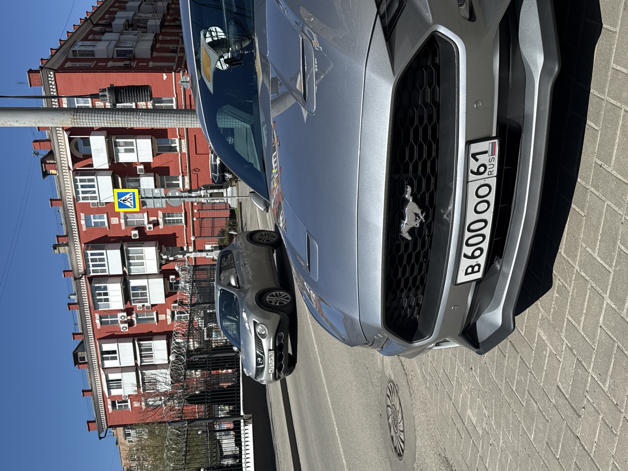 The image captures a street scene in Rostov-on-Don, Russia. A silver Ford Mustang is parked on a paved surface, its front grille and Russian license plate (B600OO 61) prominently displayed. Behind the Mustang, another car, a silver Nissan Juke with the license plate M757CP 61, is visible. The background features a tall, red brick building with white-framed windows and numerous air conditioning units. A pedestrian crossing sign (blue square with a walking figure) is positioned beside a lamppost near the building. Barbed wire stretches along a fence separating the cars from the building. The sky is blue.