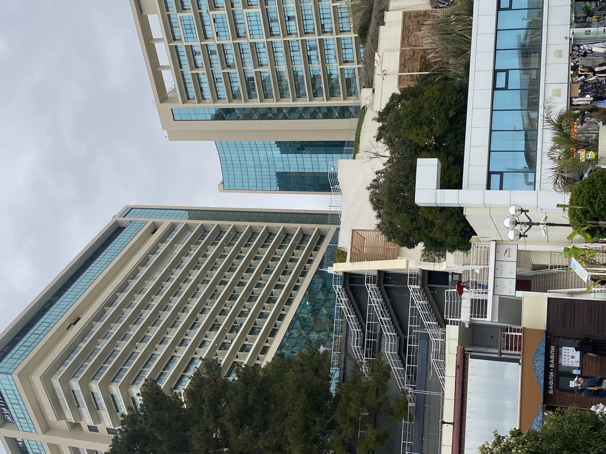 The image captures a street scene in Sochi, Russia, showcasing a blend of modern architecture and commercial activity. Two towering buildings dominate the view, each featuring a facade with numerous balconies and glass accents. Between these buildings, a smaller structure with a blue glass wall is visible.
On the right side of the image, a multi-level structure with white railings and stairs is partially obscured by trees. Adjacent to this, there are storefronts, including a cafe with a sign in Russian (ВАФЛИ • ВАФЛИ), indicating "Waffles • Waffles." People are visible near the cafe, suggesting pedestrian activity. The sky is overcast, casting a muted light over the scene.
