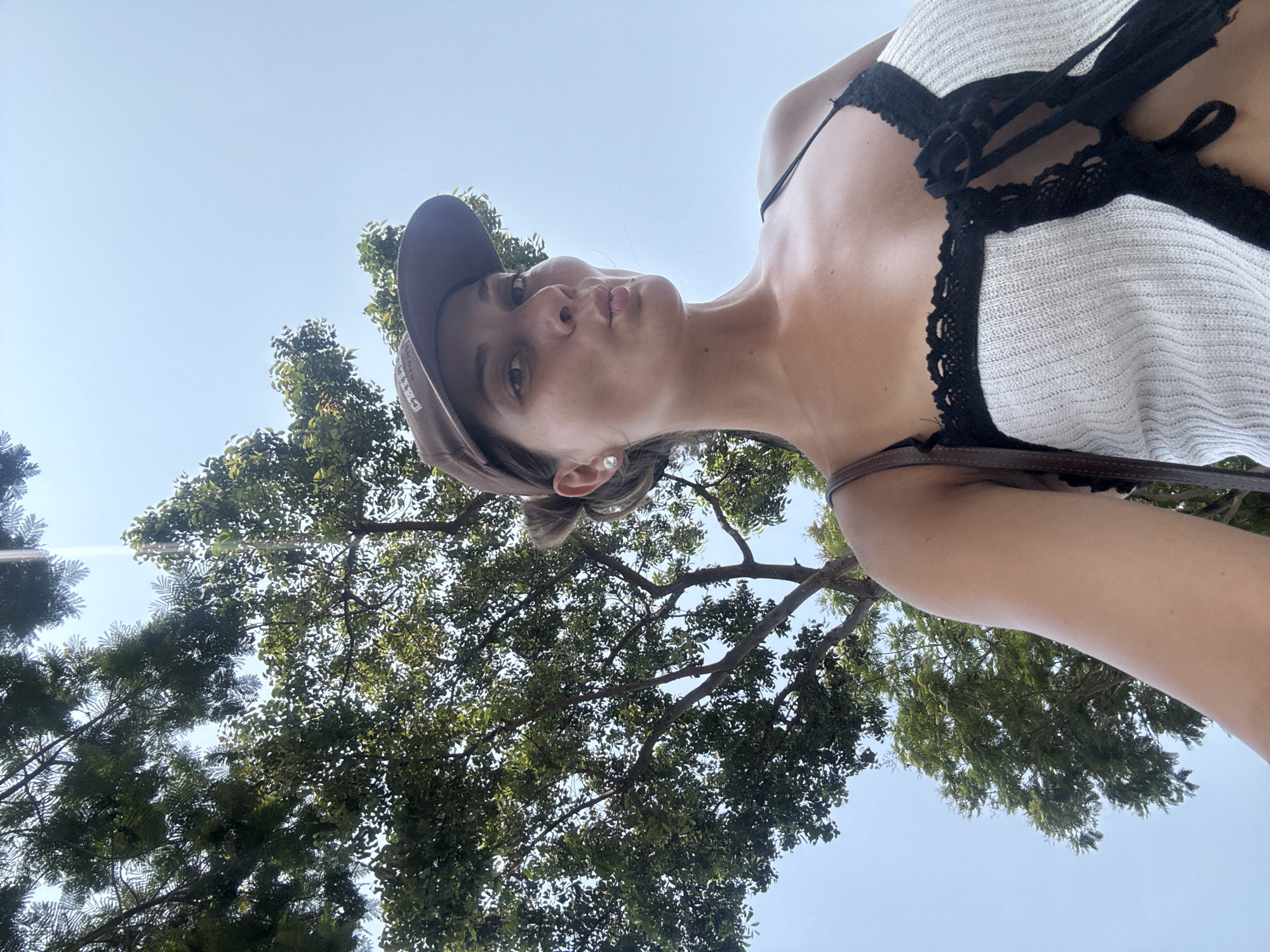 A woman wearing a brown baseball cap and a white top with black lace trim is photographed from a low angle under a large, leafy tree. The sky is visible in the background. The photograph was likely taken outdoors on a sunny day. Đà Nẵng, Vietnam is not discernable in the image.