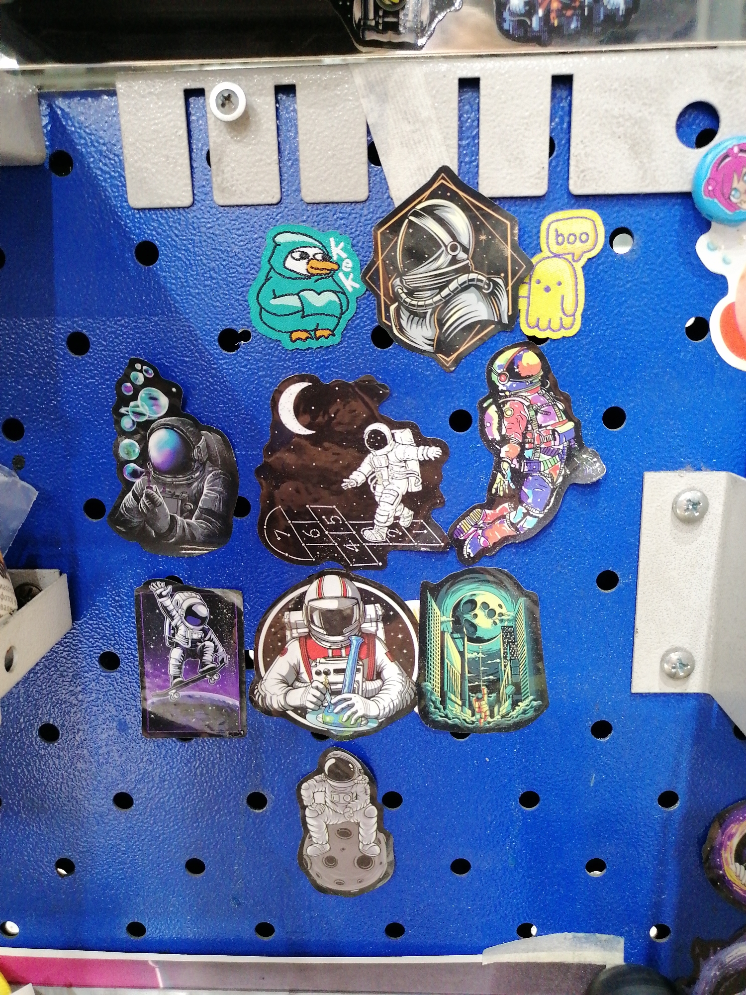 A blue pegboard is adorned with various stickers. The stickers include several astronauts in different poses: one blowing bubbles, one doing hopscotch, one skateboarding, and one sitting on the moon. Other stickers feature a penguin, a ghost, and a cityscape. The pegboard has metal supports attached with screws.