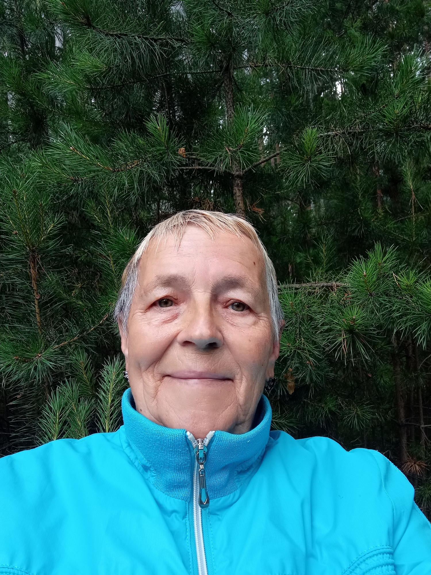 A medium shot taken outdoors shows an elderly woman with short, light-colored hair and fair skin, wearing a bright blue zippered jacket. She is smiling gently and looking towards the camera. Behind her, a dense background of dark green pine branches fills the frame. The image appears to have been taken in a natural setting, possibly a park or forest. There is no discernible indication of the specific location within Degtyarsk, Russia.