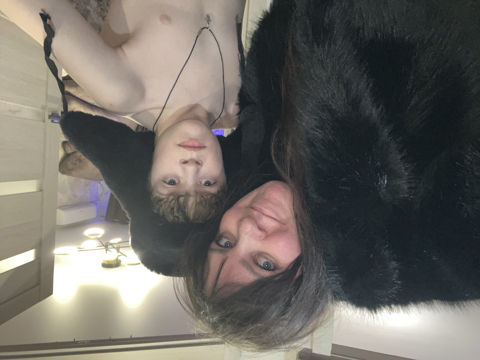 A selfie-style image depicts two individuals, one older woman and one younger person, within an interior setting. The woman, wearing a dark fur coat or similar garment, is closer to the camera and slightly below the other person. The younger individual is fair-skinned, shirtless, and wears a necklace. The background includes a glimpse of a chandelier-style light fixture and a section of wall with vertical slats. The image orientation is rotated. There is no information to suggest the location being in Noviy, Russia.