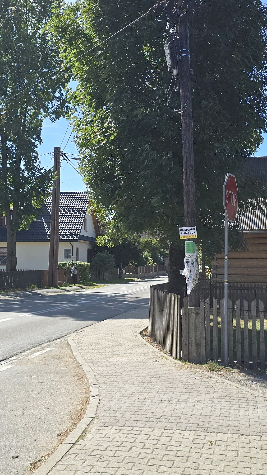 The image shows a paved sidewalk curving alongside a road in Chochołów, Poland. A wooden fence runs along the right side of the sidewalk. A utility pole stands near the fence, adorned with several flyers and a stop sign. In the background, a building with a dark, tiled roof is visible, as are trees and other houses. A person is walking on the sidewalk in the distance. The environment appears to be a residential or semi-rural area on a clear day.