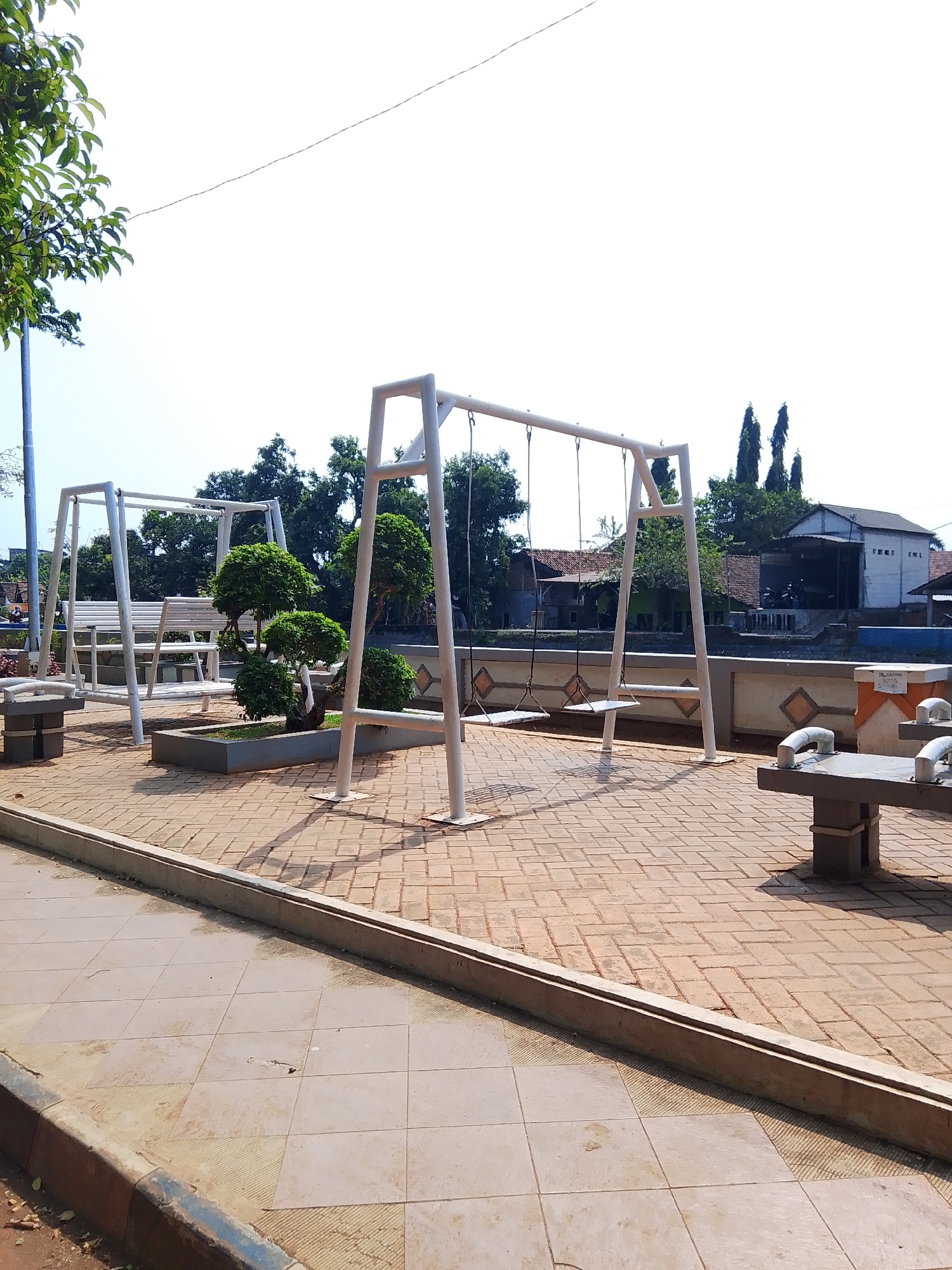 The image depicts a public park area with two white metal swing sets. One swing set, on the left, has a bench attached. The ground is paved with brick-like tiles. In the center of the image, there is a planter with several small, manicured trees. Behind the swings, a wall separates the park from a body of water, with houses and trees visible on the opposite side. The sky is clear and bright. The scene appears to be located in Potroyudan, Indonesia, as indicated by the information provided. There are no people visible in the image.