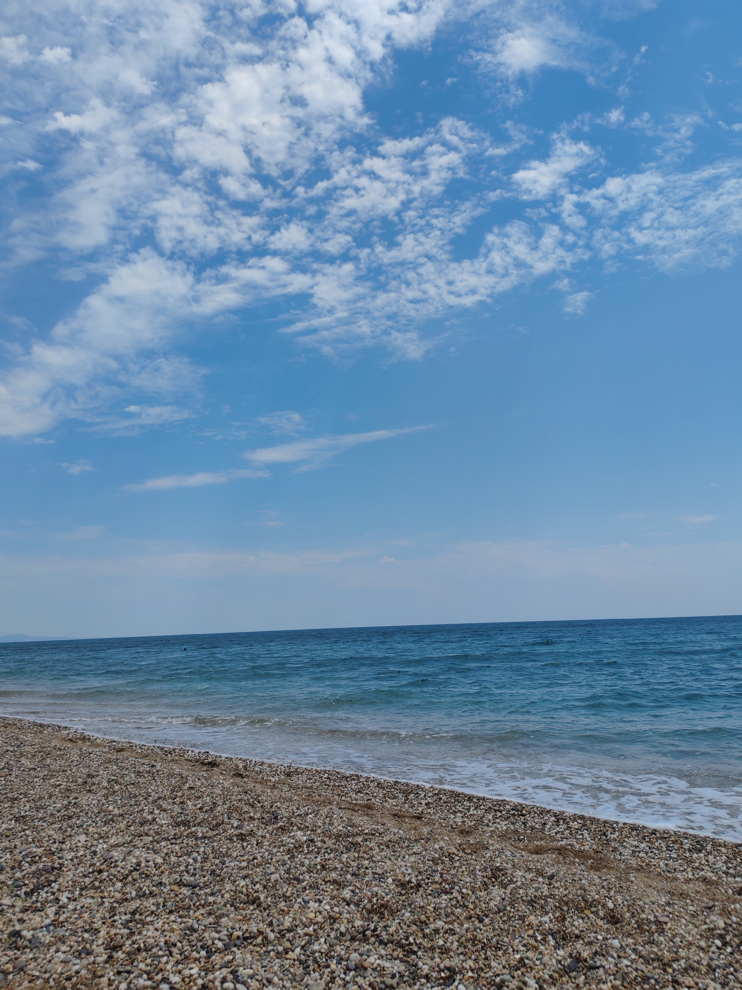 This media file depicts a tranquil beach scene in Makri, Greece. The image is dominated by a clear blue sky with scattered white clouds, suggesting fair weather. The foreground is a pebble beach with a mix of light and dark stones. Gentle waves from the deep blue Aegean Sea lap at the shore, creating a white foamy line. The sea extends to the horizon, where a faint outline of distant land can be seen. There are no people or objects visible on the beach or in the water. The overall impression is one of peace and natural beauty, likely captured during the daytime given the bright lighting.