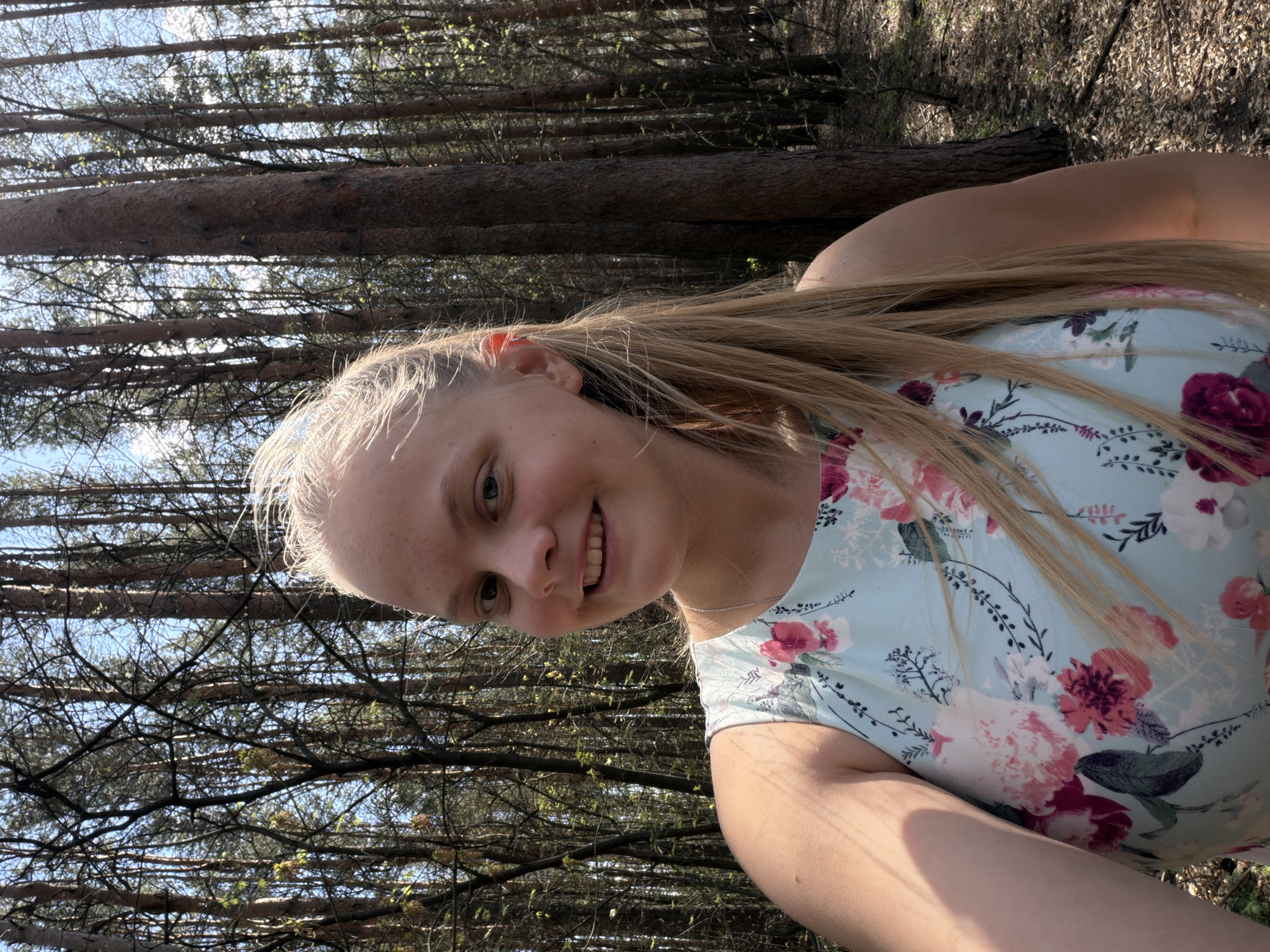 A young woman with blonde hair smiles at the camera in an outdoor setting. She is wearing a light blue floral dress. The background consists of a forest with tall trees. The location is not specified, but the request mentions Izhevsk, Russia.