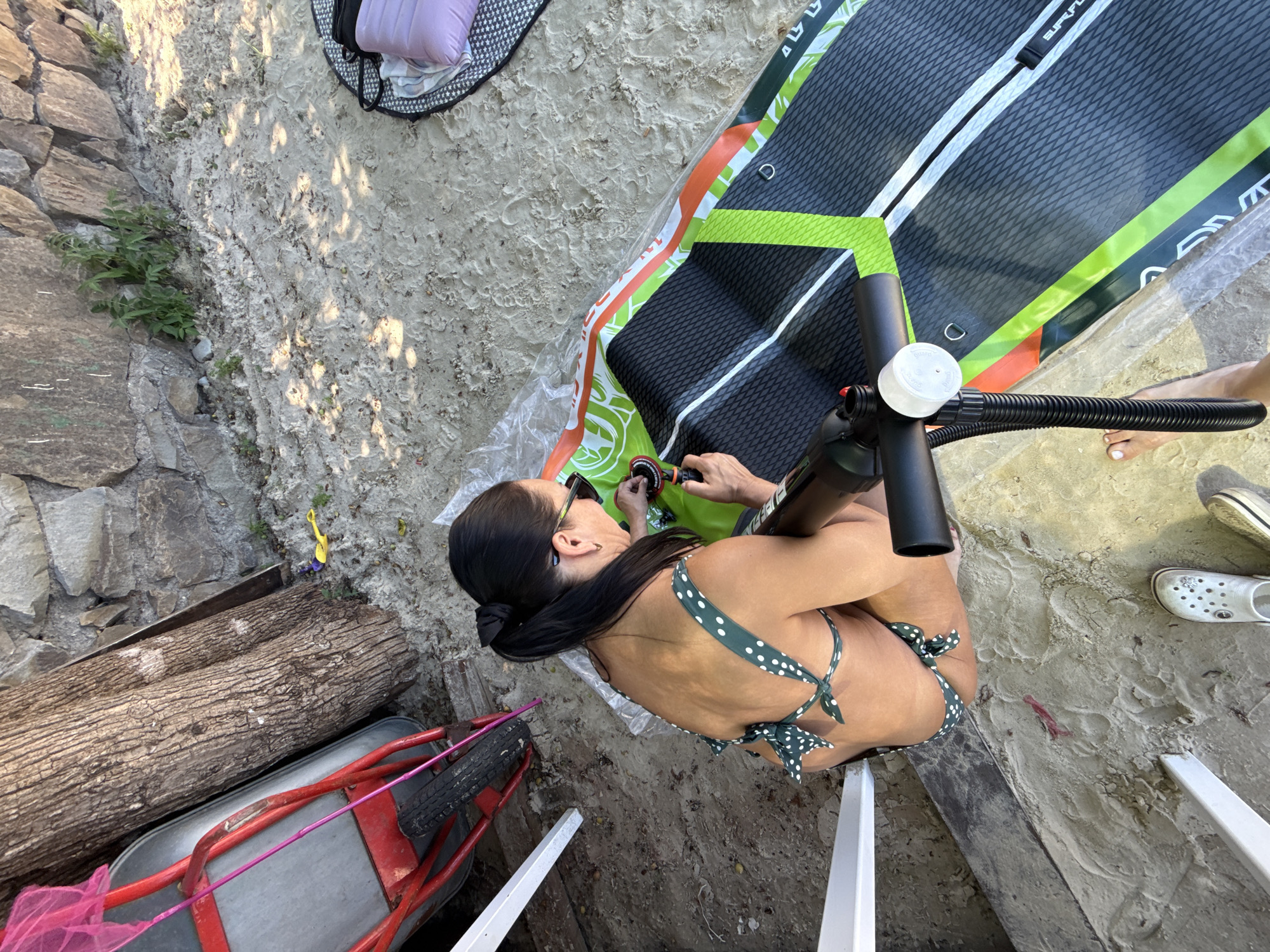 In this overhead shot, a woman in a green polka-dot bikini is on a sandy beach, preparing a paddleboard. The paddleboard is vibrant with green, white, and orange accents, and features the text "SURF PLUS" along its side. The woman is actively engaged with a black and silver pump, its hose connected to the paddleboard. Her posture suggests focus as she works on inflating the board.
The scene is set on a beach with a rocky embankment on the left, and scattered logs and a wheelbarrow in the foreground. The sand appears dry and marked with footprints, indicating it is likely a sunny day. Shadows cast by an unseen object suggest it is either morning or late afternoon. The overall mood is one of outdoor leisure and preparation for an activity. The woman wears sunglasses, further hinting at bright conditions. Two white Crocs are visible near the paddleboard, suggesting the presence of another person or preparation for more than one person.