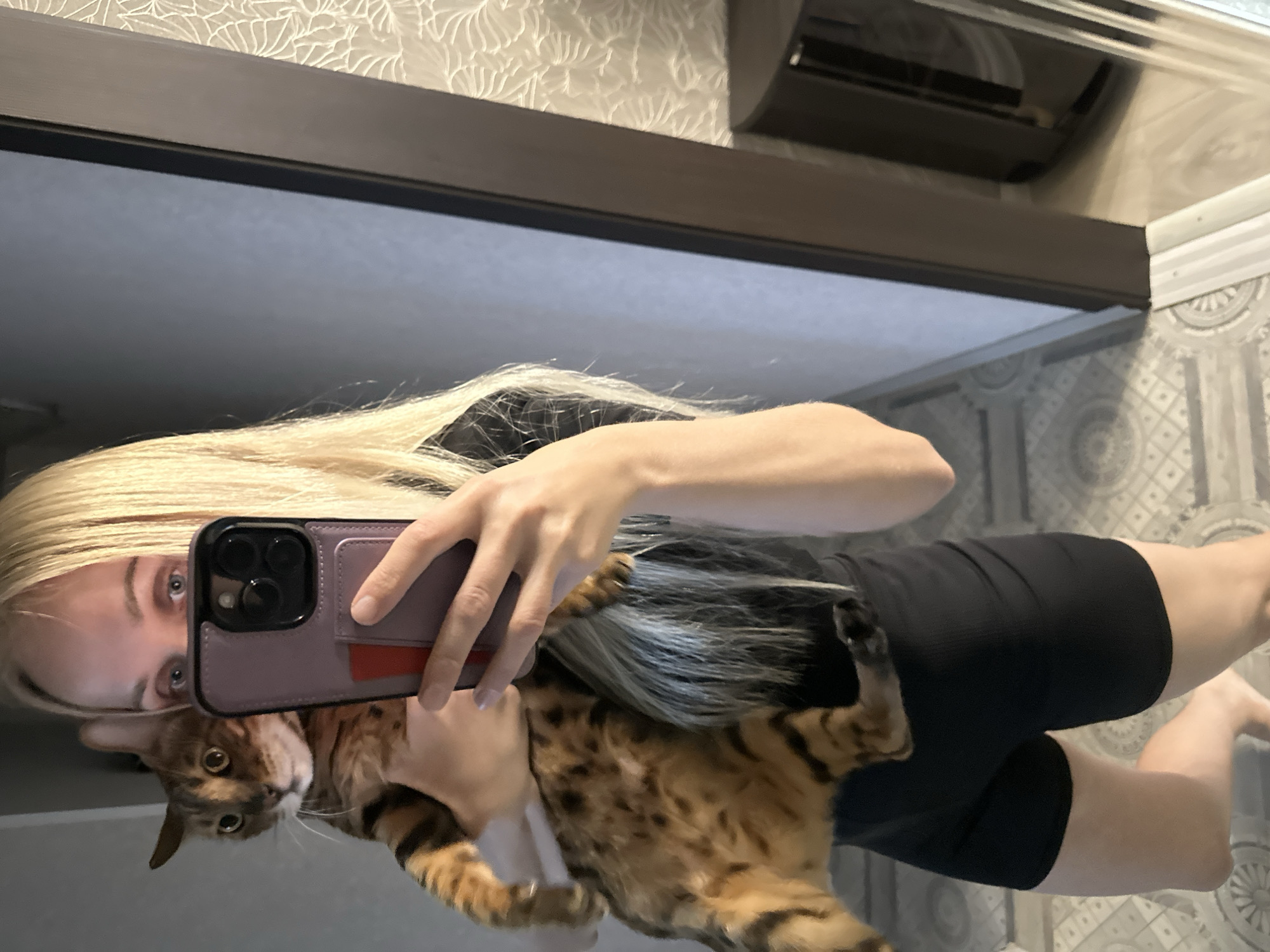 A person with blonde hair is holding a Bengal cat and taking a selfie in a mirror. The person is wearing black shorts and a black top. The cat is looking at the camera. The background shows a wall with wallpaper and a floor with a patterned tile. The image appears to be taken indoors, possibly in a home. The city of Energetik, Russia is not depicted in the image.