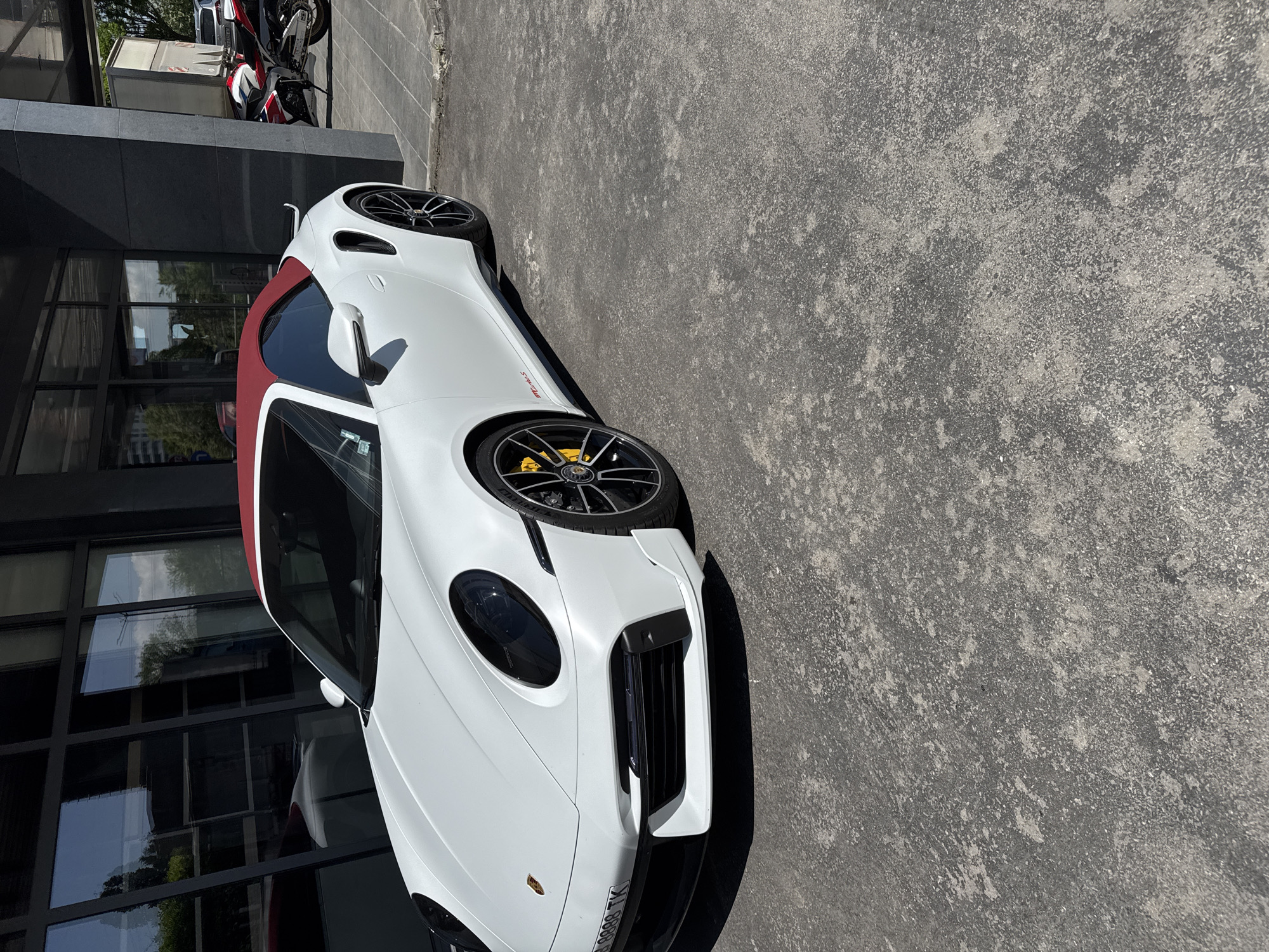 The image shows a white Porsche 911 GT3 RS parked on a gray concrete surface next to a building with glass windows. The car has a red roof and black wheels with yellow brake calipers. In the background, a red and white motorcycle is visible next to the building. The license plate of the car is visible. The photo was taken in Sofia, Bulgaria.