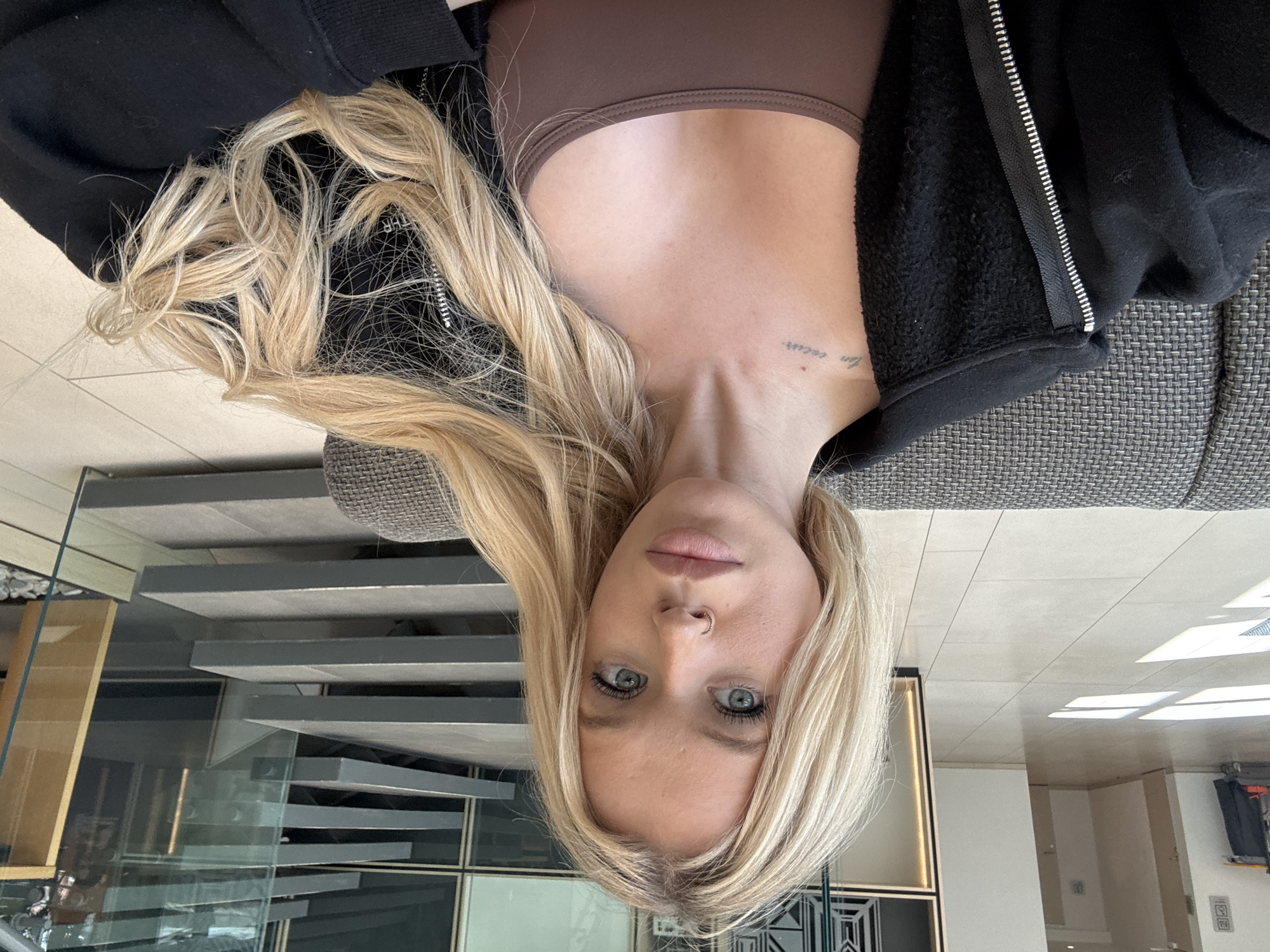 This is a horizontal, slightly tilted, close-up selfie of a young Caucasian woman with long, blonde hair and blue eyes. She is lying down, with her head tilted to the side, and looking directly at the camera. She is wearing a brown bra and a black zip-up hoodie. A small, faint tattoo is visible on her neck, reading "mon coeur." Her nose piercing is also visible. The background consists of a sofa with a textured grey fabric, a modern staircase with glass railings, and some wooden furniture. The lighting suggests it is daytime, with natural light coming from the right. The overall mood is relaxed and casual. No weather cues are apparent. The setting appears to be an indoor, contemporary living space.