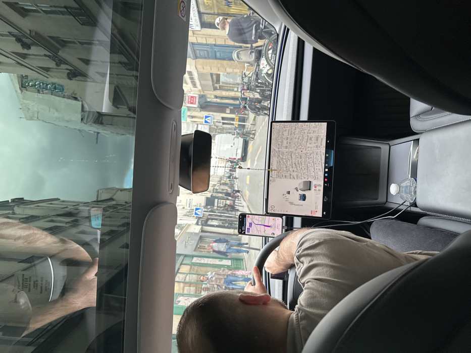 The scene depicts a street view from inside a moving vehicle, likely a Tesla due to its distinctive interior features. A male driver, seen from the rear, with short hair and wearing a light-colored t-shirt, has both hands on the steering wheel. A smartphone displaying a navigation application with a purple route is mounted vertically to the left of the car's large central touchscreen, which shows a city map and a driving visualization indicating surrounding vehicles. A rosary bead chain hangs from the rearview mirror assembly. A small, clear plastic water bottle rests in the center console.
The vehicle is traveling down a narrow, bustling street in Paris, France, characterized by multi-story, light-colored stone buildings with classic Parisian architecture. Many buildings feature ground-floor storefronts. Pedestrians are visible on both sidewalks and crossing the street. A significant number of bicycles are parked along the right-hand sidewalk, along with at least one scooter. Further down the street, a white delivery van is visible. Scaffolding is present on the upper portion of a building on the right side of the street. The sky overhead is overcast. Reflections in the car's glass roof include an arm and text that appears to read "NAPOLEON DETOUR," suggesting proximity to a related landmark or a temporary diversion sign in the city.