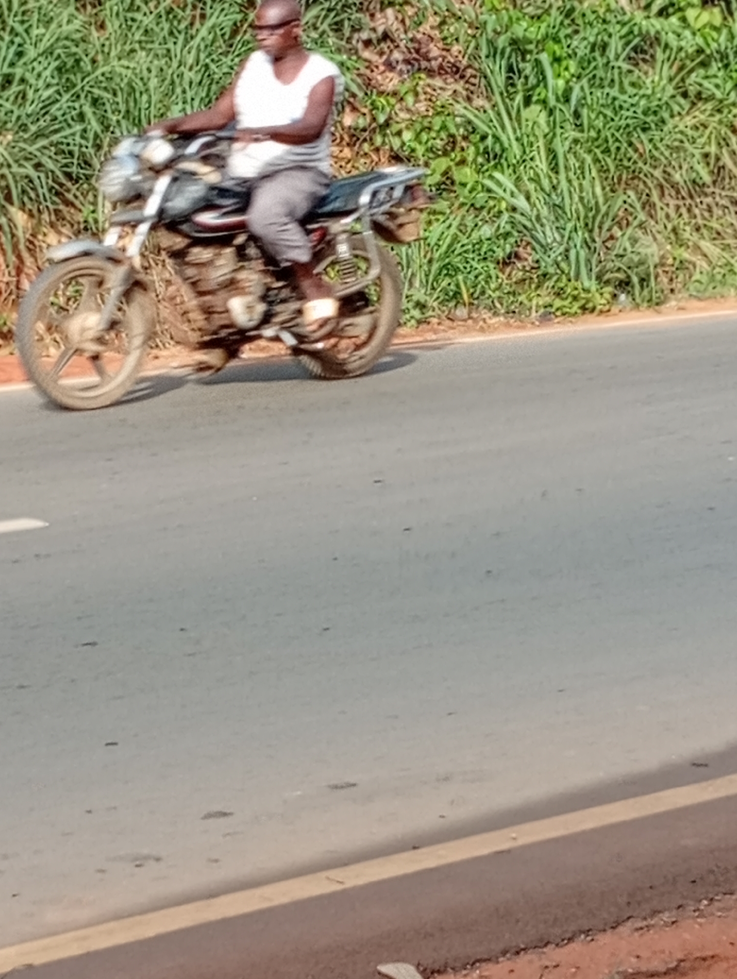A man rides a motorcycle on a paved road in Nkpor, Nigeria. He is bald, wearing glasses, a white t-shirt, and grey shorts. The motorcycle is grey and black. Lush green vegetation grows along the roadside.