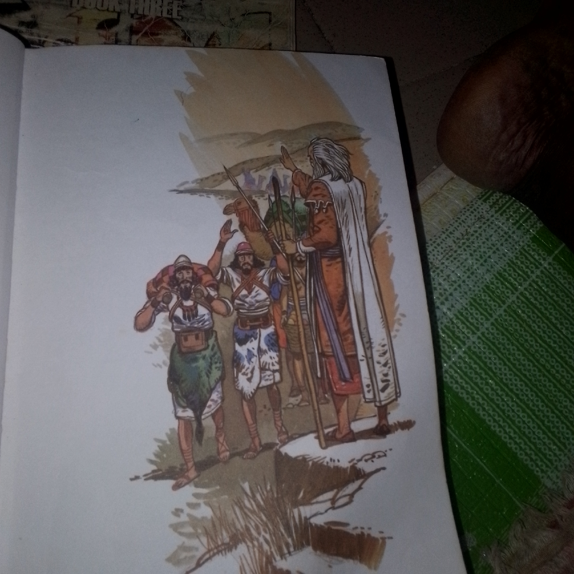 This media file shows a watercolor illustration from a book. The illustration depicts a group of people in what appears to be an arid, desert-like landscape. In the foreground, three men are visible. The man on the left is carrying a bundle on his shoulders. He is wearing a helmet, a tunic with a chest strap, and a green skirt-like garment. The man in the center is also wearing a helmet and a patterned tunic, and he is raising his hand. He appears to be carrying a spear. Behind him, a camel is partially visible. To the right, a taller figure in an orange robe and a white cloak stands, also with his hand raised. This figure is leading the group. The background features a sandy, hilly terrain under a pale sky, suggesting a desert environment. The overall mood of the illustration is one of travel or procession. There is no visible text in the illustration itself, but the hint image shows "BOOK THREE" printed on the book's cover behind the illustration. The scene evokes a sense of ancient or biblical times. The time of day appears to be daytime, possibly late afternoon given the warm tones of the landscape. There are no clear indicators of weather, though the arid setting implies dry conditions.