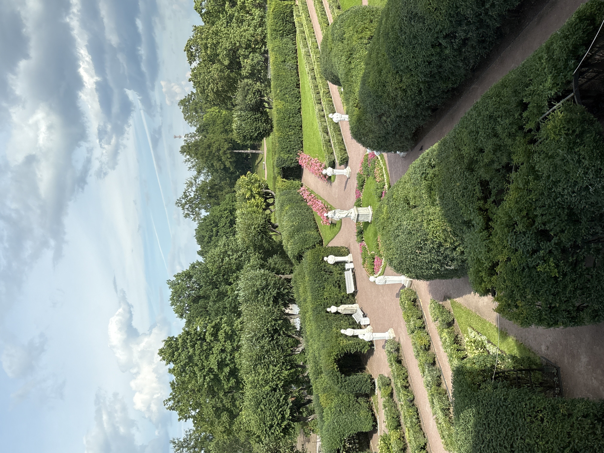 The image depicts a meticulously maintained formal garden with sculpted hedges, gravel pathways, and classical statues. The garden features manicured green hedges forming geometric patterns and archways. Several white statues of figures in classical attire are placed at intervals along the paths. Flowerbeds with pink flowers are visible in some areas. The pathways are a reddish-brown color, typical of crushed gravel. In the background, mature trees create a lush green backdrop, and the sky above is partly cloudy with hints of blue. This scene is characteristic of the gardens at the Gatchina Palace, located in Gatchina, Russia.
