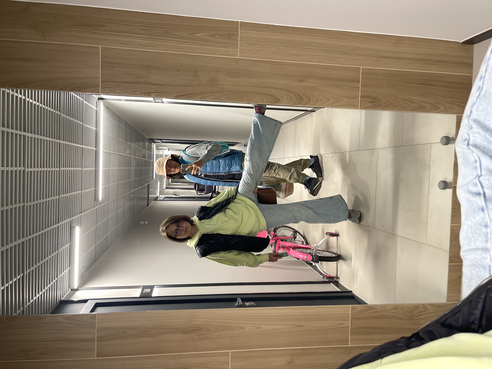 The image shows a reflection in a mirror of two people standing in a corridor. The corridor is lit by fluorescent lights, and has a tiled floor. The first person is standing closest to the mirror holding a pink bicycle. They are wearing glasses, light blue jeans, and a light green top. The second person is standing further back in the corridor, holding a phone in their right hand. They are wearing a brown hat, a light blue jacket, and tan trousers. The corridor appears to be in a residential or commercial building.