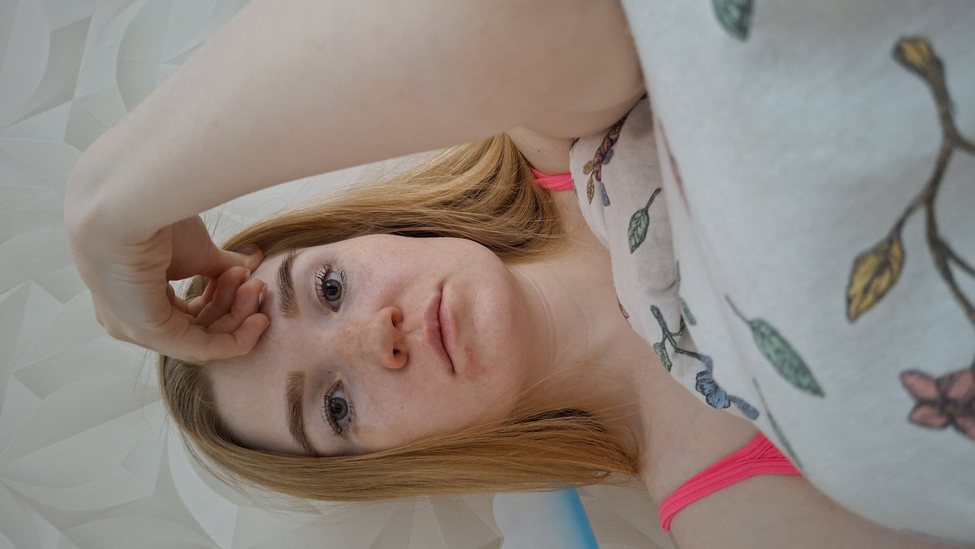 A fair-skinned young woman with freckles and reddish-blonde hair is lying on her back on a white surface with a geometric pattern, possibly a bed or floor. She is wearing a light-colored top with a floral pattern, partially obscured by her shoulder and arm. A bright pink strap, potentially from an undergarment or bathing suit, is visible at her neck. Her left hand is raised towards her forehead, with fingers touching her face. She is looking directly at the camera. There is no information to determine if this image was taken in the city of Chaikovsky, Russia.
