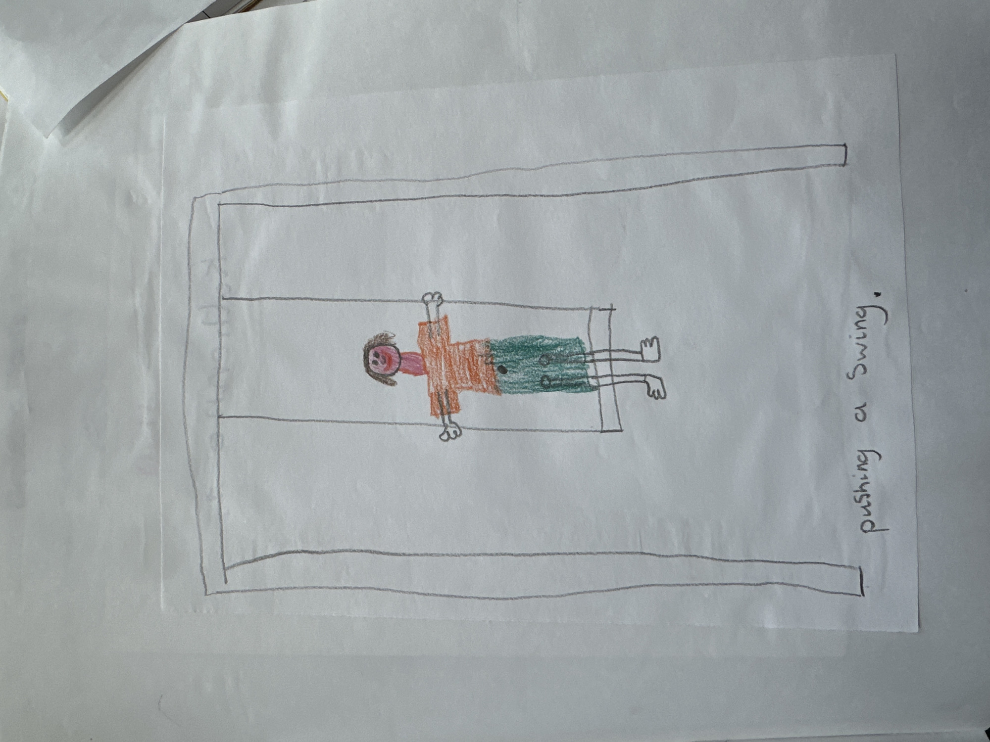 The image, originating from Wealden, United Kingdom, displays a child's drawing rendered on a white sheet of paper. The central subject is a simplified human figure positioned on a swing within a grey-lined swing set frame. The figure possesses a pink head with brown hair, black circular eyes, and a red line mouth. Its torso is an orange, horizontally striped shirt, from which two outstretched horizontal arms extend, terminating in rudimentary hands. The lower body is clad in green pants with small black dots, and two simplified feet are visible. The swing set comprises two vertical grey posts, a horizontal grey crossbar, and two grey suspension lines attaching to a grey swing seat. Below the drawing, handwritten in black ink, are the words "pushing a Swing." The surrounding environment is the plain white surface of the paper, with other sheets faintly visible in the background of the overall photograph.