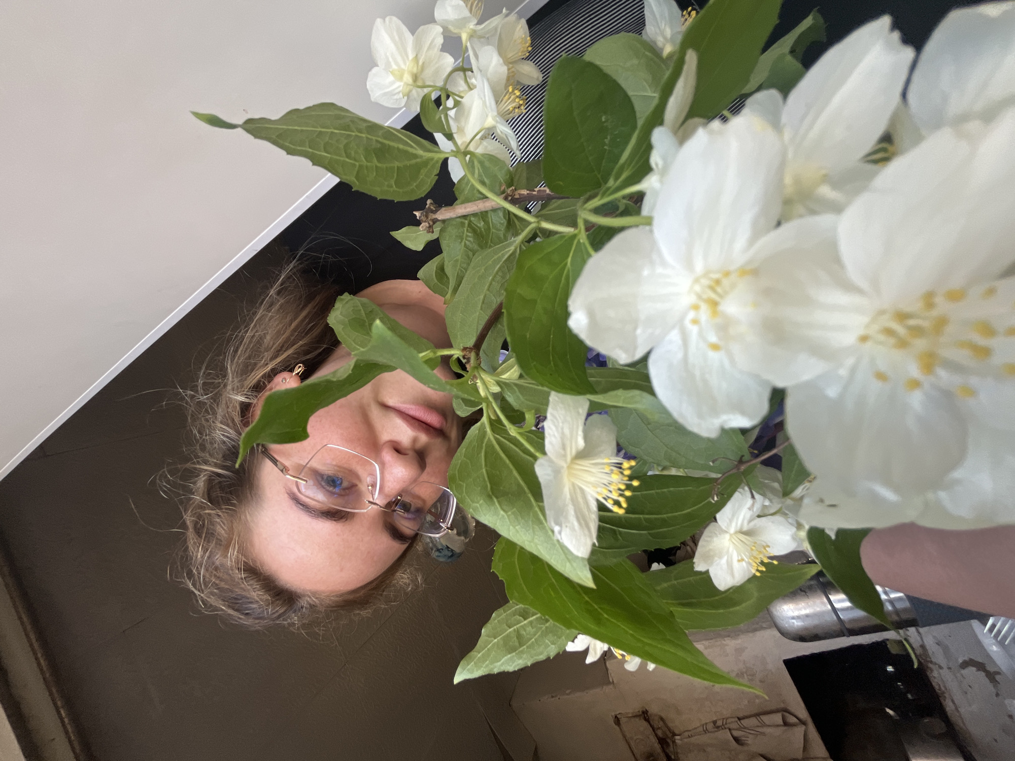 A young woman with light-colored, curly hair and rimless eyeglasses holds a bouquet of white flowers with prominent yellow stamens and green leaves. She looks towards the camera with a slight smile, her head tilted. Her left hand is partially visible, supporting the flowers. The background on the left features an indoor wall with a rough, light-brown texture, a dark rectangular opening, and a metallic cylindrical object, along with a hanging cloth. The background on the right consists of a bright, plain white wall or ceiling, contrasted by a dark, angled surface below it. The scene takes place indoors in the city of Sosnovo, Russia.