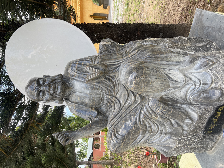 A dark grey stone statue, veined with lighter grey and gold, depicts a seated elderly male figure with a wrinkled face, prominent ears, and an open mouth, wearing draped robes. The figure's left hand is raised with the thumb and forefinger touching, while the right hand is cupped, holding an open book or scroll. A large, flat, circular white disc is positioned directly behind the statue's head. The statue is situated on a rough-hewn stone base, which rests upon a dark grey polished rectangular plinth inscribed with "TÔ PHỤC-ĐA-MẠT-ĐA".
The scene is an outdoor area within a temple complex located in Đăk Mar, Vietnam. Dense green coniferous tree branches extend into the upper left and right portions of the frame, partially obscuring the white disc and background. To the right of the statue, a multi-story building with yellow walls and arched openings, consistent with temple architecture, is visible. To the left, a building with a red roof and reddish-brown walls is partially visible through intervening trees. The ground consists of sparse grass and light-colored earth. A small potted plant with red blossoms is positioned to the left of the statue's base.