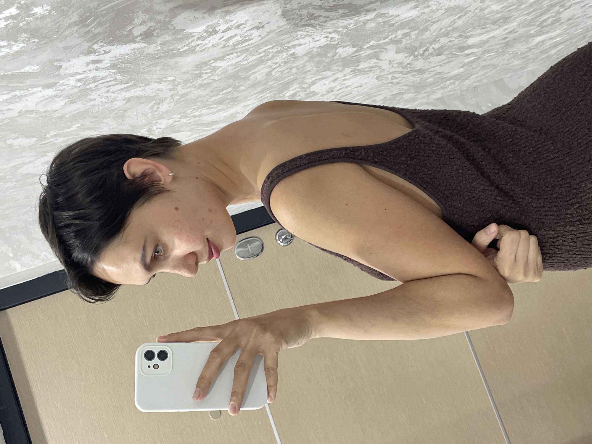 A woman with short, dark hair and fair skin is taking a selfie with a white smartphone. She is wearing a brown, textured dress with thin straps. Her left arm is extended, holding the phone, while her right arm is bent and her hand is near her shoulder. She is looking down at the phone screen. The background is a vertically oriented wall with what appears to be a light beige wall with a textured white surface above. The lighting is bright and appears to be from an artificial source. The location is not identifiable as Izhevsk, Russia based on the visual information provided.