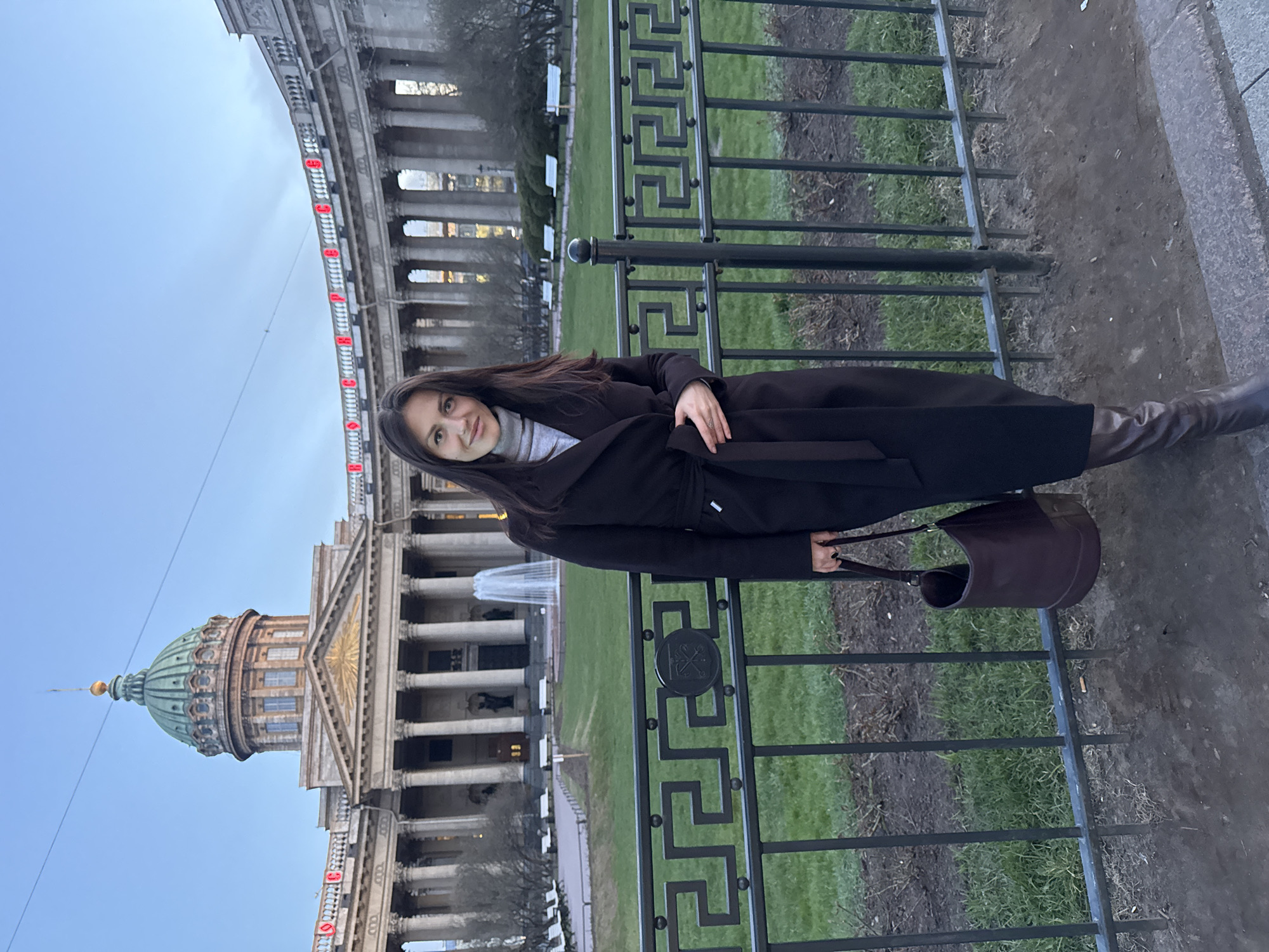 A woman wearing a black coat and brown boots poses in front of the Kazan Cathedral in Saint Petersburg, Russia. She is standing near a black metal fence and holding a brown handbag. The Kazan Cathedral, with its distinctive green dome and colonnade, is visible in the background. The sky is overcast.