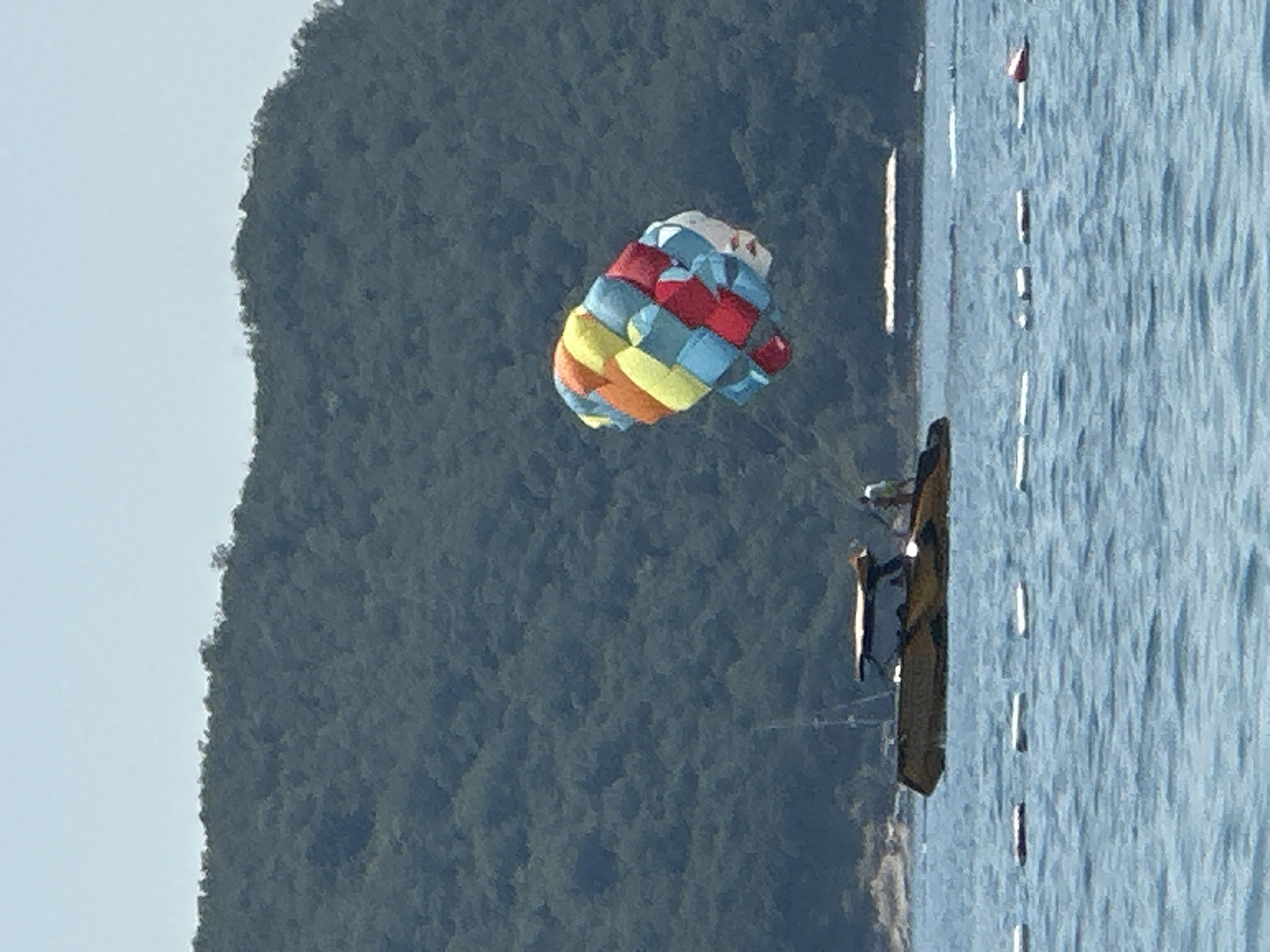 The image captures a parasailing activity on a bright, sunny day in Boreti, Montenegro. A colorful parasail, a patchwork of red, blue, yellow, and orange squares, is deployed in the sky above a calm, blue body of water. The parasail appears to be pulled by a boat, which is partially visible in the lower right of the frame, a long, dark yellow vessel with a person sitting on it. The boat is moving through the water, creating subtle ripples.
In the background, a densely forested hillside rises steeply from the water's edge, its dark green foliage creating a lush backdrop. The sky is a clear, pale blue, suggesting good weather conditions. The overall scene conveys a sense of leisure and recreation. There is no visible text in the image. The time of day appears to be mid-morning or afternoon, given the brightness and angle of the light.