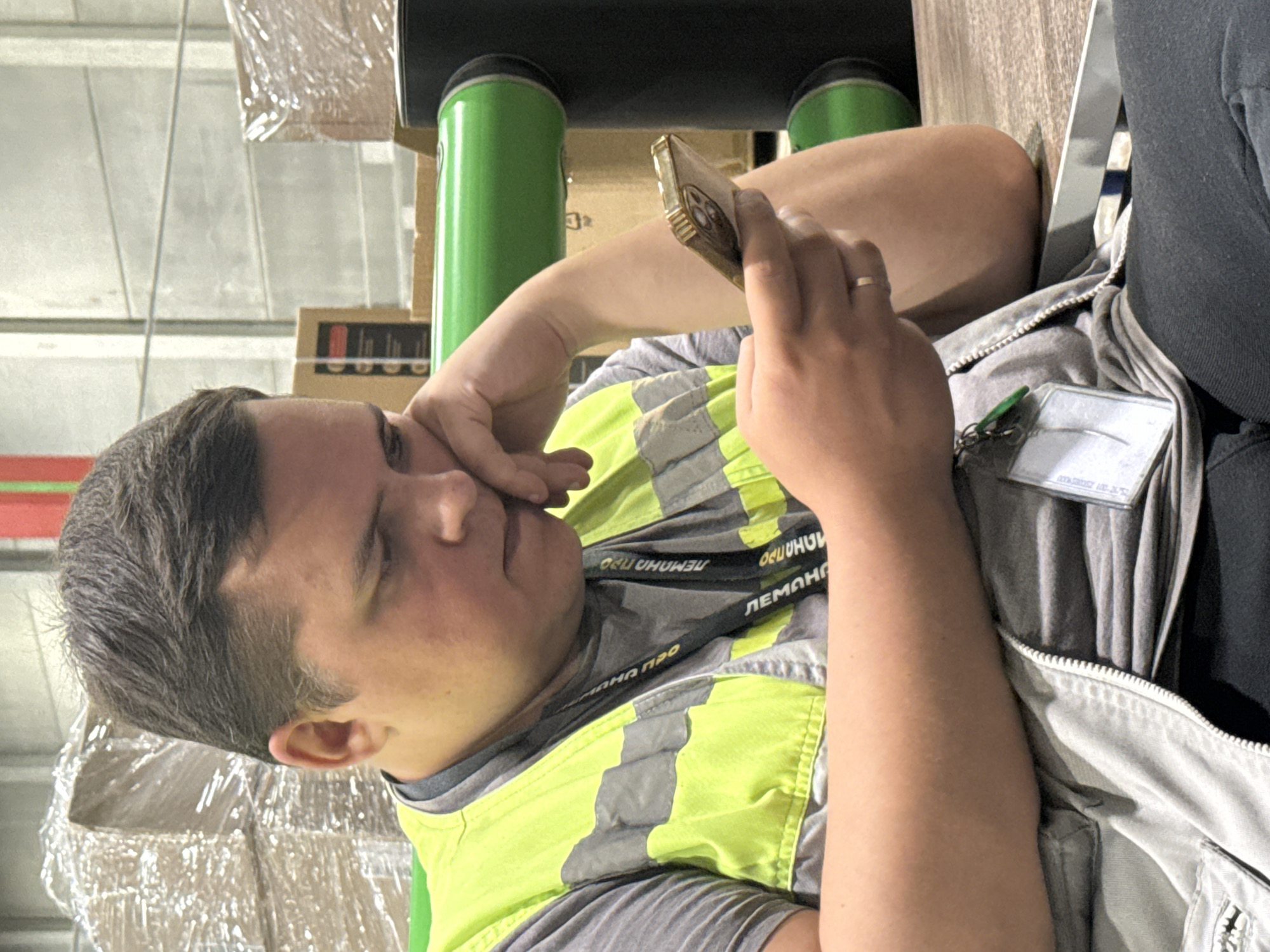 A man in a reflective vest is sitting at a table and looking at his phone. He is leaning his head on his hand. In the background, there are shelves with boxes.