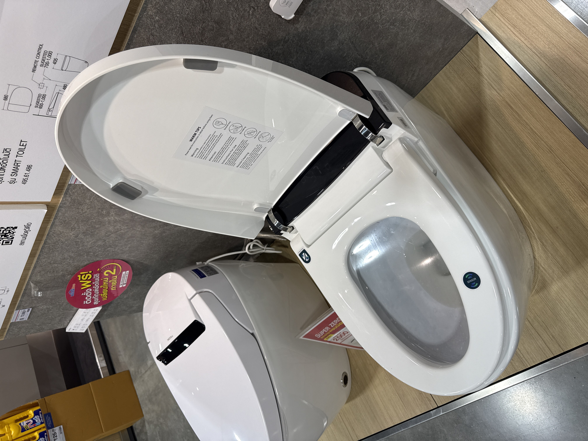 The image shows a display of modern, white smart toilets in a retail setting, likely a home improvement store. The primary focus is on the opened toilet seat of one unit, revealing the inner workings and a label with "WARM TIPS" and instructions. A second, closed toilet is visible to the left, and other promotional materials are present on the wall behind the toilets, including price tags and diagrams. The flooring appears to be wood-look tile. The scene is well-lit and appears to be an indoor environment. There are no people visible in the image. The specific location "Baan Chaweng Noi, Thailand" is not depicted or implied by the visual content of the image itself.