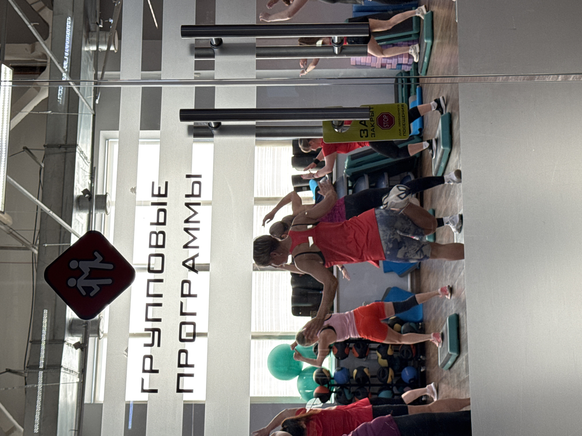 This is a photograph of people participating in a group fitness class in a gym. The image is taken from a slightly elevated angle, looking down at the participants and the gym environment. The lighting suggests it is daytime.
The primary subjects are a group of women dressed in athletic wear, engaged in what appears to be a high-intensity interval training or aerobics class. They are positioned on and around aerobic step platforms. Some are performing exercises, while others are in various stages of movement.
Visible objects include green and blue aerobic step platforms, various weights and fitness balls in the background, and gym equipment like metal racks and ventilation systems. The gym has large windows that allow natural light to filter in, and the floor appears to be wood.
The text "ГРУППОВЫЕ ПРОГРАММЫ" (Group Programs) is prominently displayed on a frosted glass panel, indicating the type of service offered. A yellow sign with red lettering reads "ЗА" (For) and "ЗАКРЫТ STOP" (Closed STOP), suggesting a temporary closure or restricted access for a specific purpose, possibly "для свободного посещения" (for free attendance) as partially visible.
The overall impression is one of an active and dynamic fitness environment. The women appear focused on their exercises. The image's orientation is rotated 90 degrees clockwise.