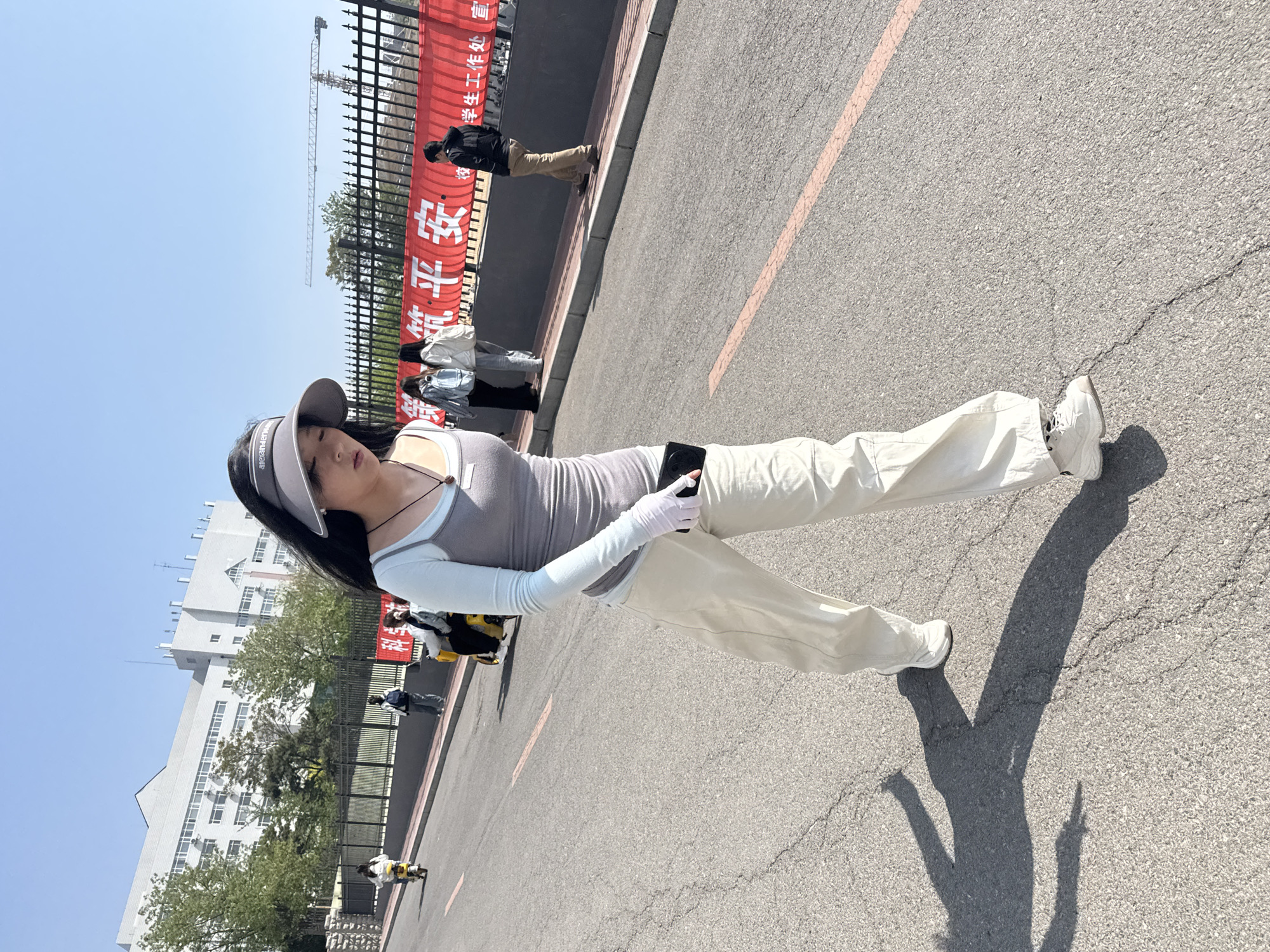 A young Asian woman in a visor, gray top, light blue long-sleeved shirt, light pants, and white sneakers stands on a gray asphalt road holding a phone in her gloved hand. Her shadow is cast on the road. In the background is a metal fence, a red banner with Chinese characters, a large white building, and trees. Several pedestrians are walking in the background. The location is likely Zhongtun, China. A crane is visible in the background.