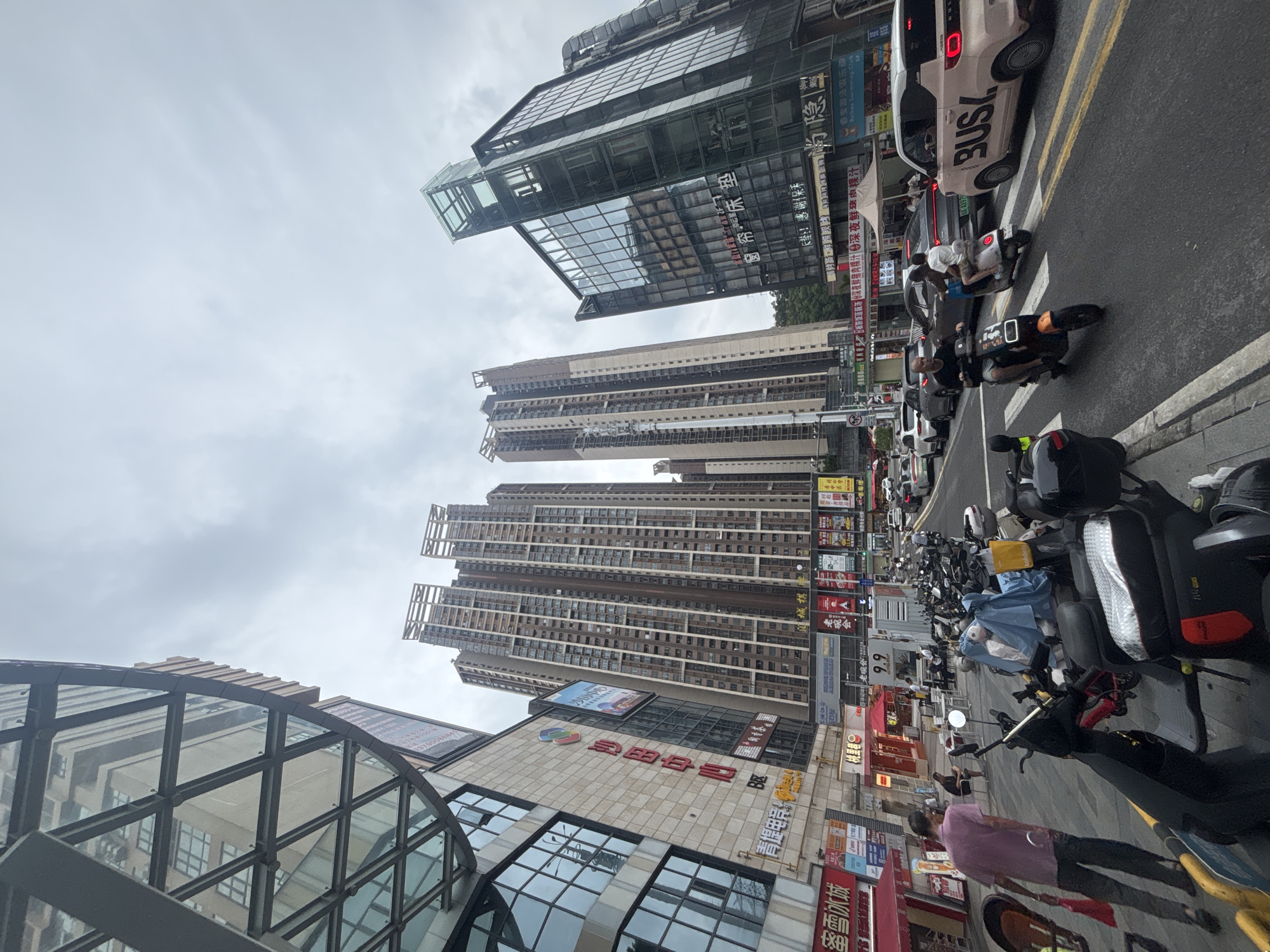 The image captures a street scene in Nanhai District, China, dominated by towering apartment buildings and modern commercial structures. In the foreground, a row of parked motorcycles and scooters lines the sidewalk. A man in a pink shirt walks away from the viewer, carrying a red bag. Across the street, vehicles, including cars and scooters, navigate the roadway. A light-colored minivan with "BUSL" written on its side is visible in the right lane. Pedestrians can be seen on the sidewalk, some sitting at tables outside shops. The architecture is a mix of glass-fronted buildings and traditional apartment blocks with numerous balconies. The sky is overcast. Advertisements and signs in Chinese characters adorn the storefronts.