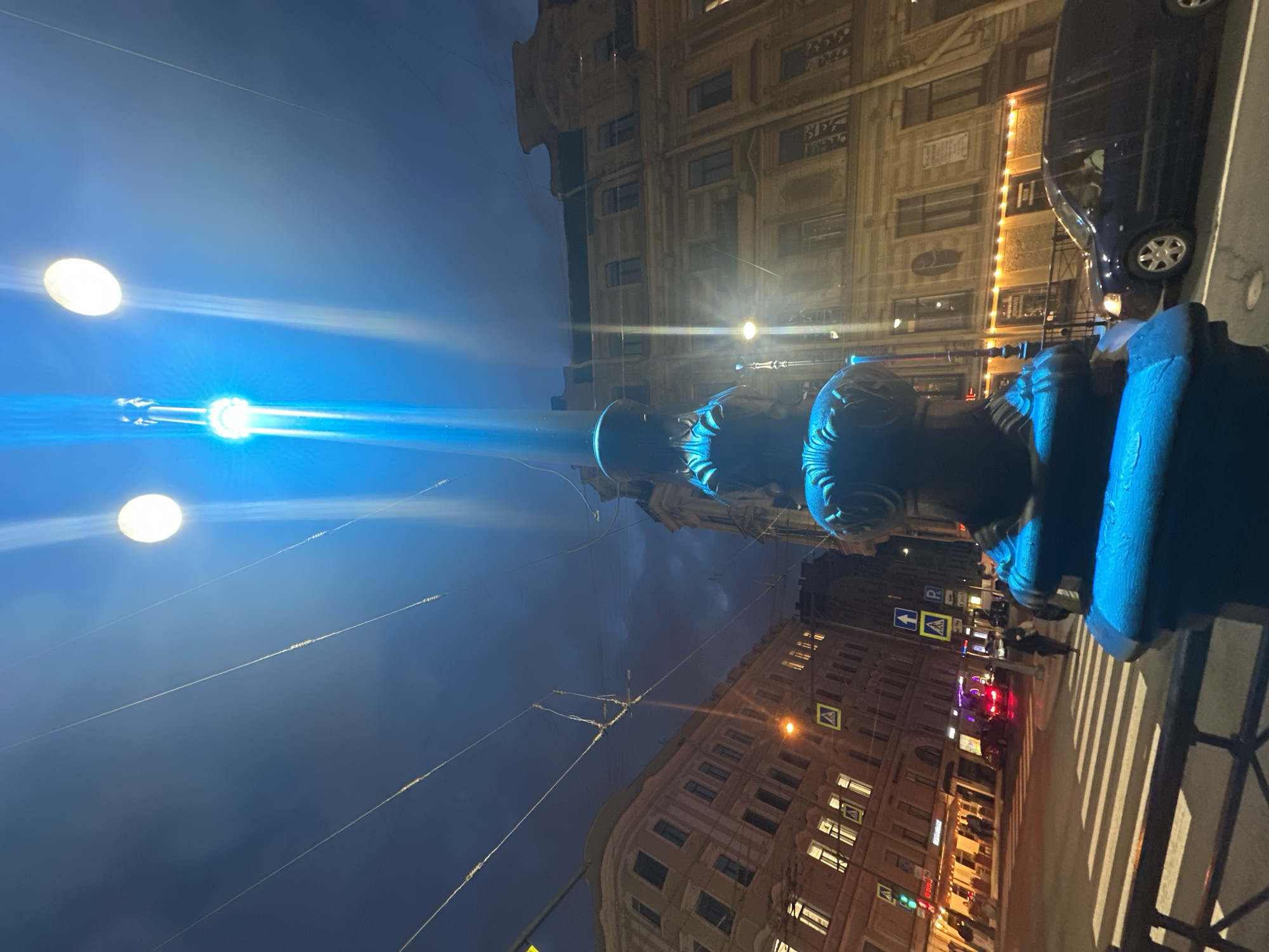 This nighttime photograph, taken in Saint Petersburg, Russia, is shot from a low angle looking upwards. The main subject is a blue-painted metal post on a street corner. The background reveals a cityscape with buildings illuminated by warm light, a dark blue sky with streetlights beaming bright blue, and power lines crisscrossing the sky. On the right side of the image, a car is parked on the street. A pedestrian crossing with white stripes is in the foreground to the right of the post. In the distance, traffic lights and other illuminated signs are visible. Buildings are on either side of the frame, lit up with lights.
