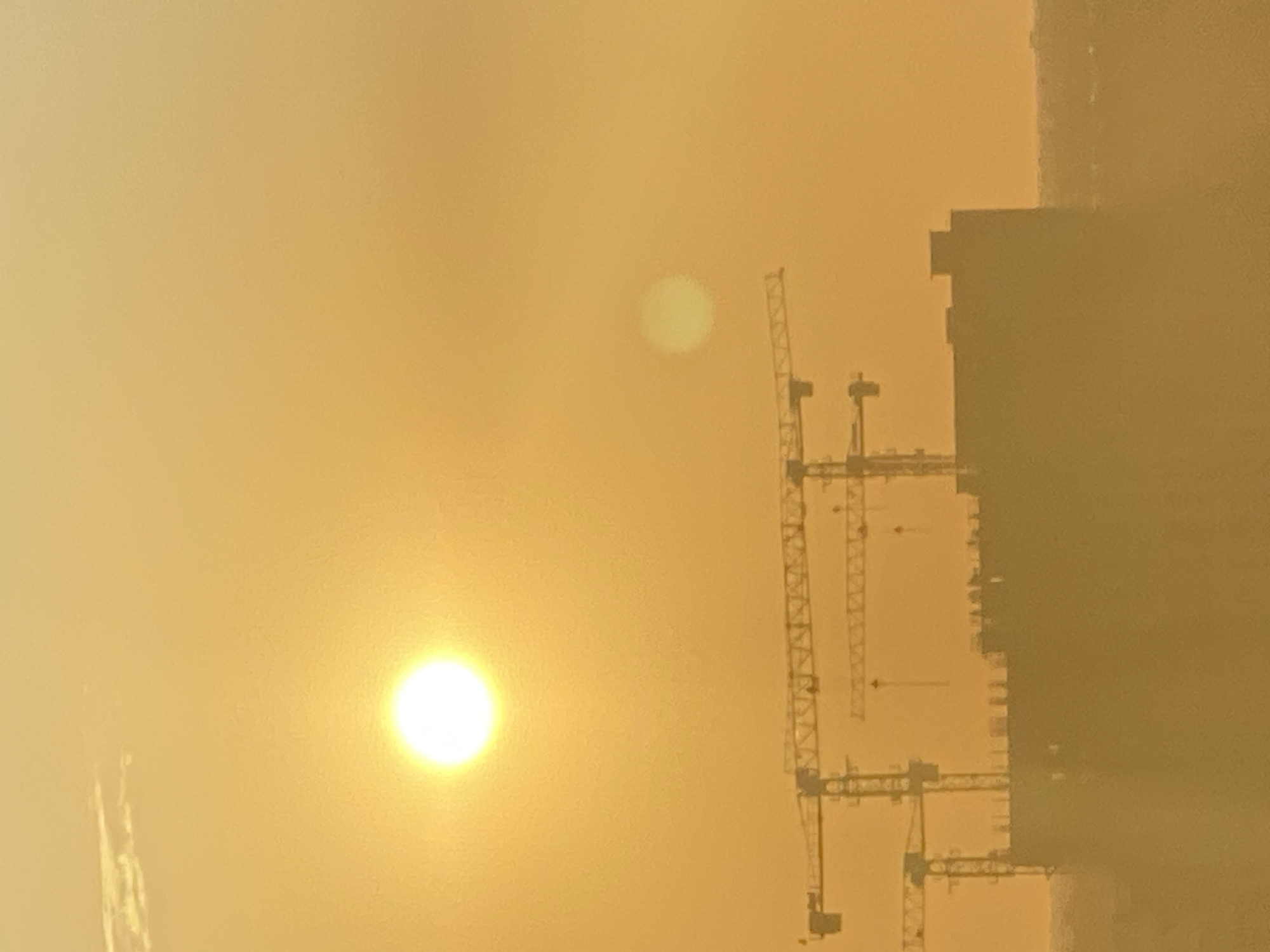 The image captures a hazy, golden-hued sunset over a city skyline. The sun, a bright, luminous orb, is prominently positioned in the upper left quadrant of the frame. To its right, a fainter, diffused disc, likely another celestial body or atmospheric phenomenon, is visible.
Dominating the mid-ground and lower half of the image are several construction cranes, silhouetted against the bright sky. Their metallic frames are intricate and skeletal, suggesting an active construction site. Beneath the cranes, the indistinct, dark mass of a partially constructed building or a dense cluster of buildings is visible. The overall impression is one of urban development under a dramatic sky.
The atmosphere appears thick with haze or dust, softening the edges of all elements and giving the scene a dreamlike quality. The colors are predominantly warm, ranging from bright yellow near the sun to muted oranges and browns in the lower portions of the image. The lack of clear details in the background suggests a distant cityscape obscured by the atmospheric conditions. There are no people visible in the frame.