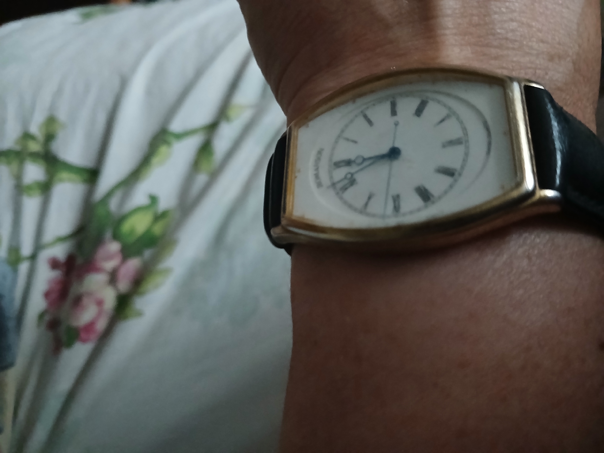 A close-up shot shows a person's arm with a gold-toned watch featuring a white face and Roman numerals. The watchband is black leather. The background is out of focus, showing a white fabric with a floral pattern in pink and green. The setting is presumed to be Hlushky, Ukraine.